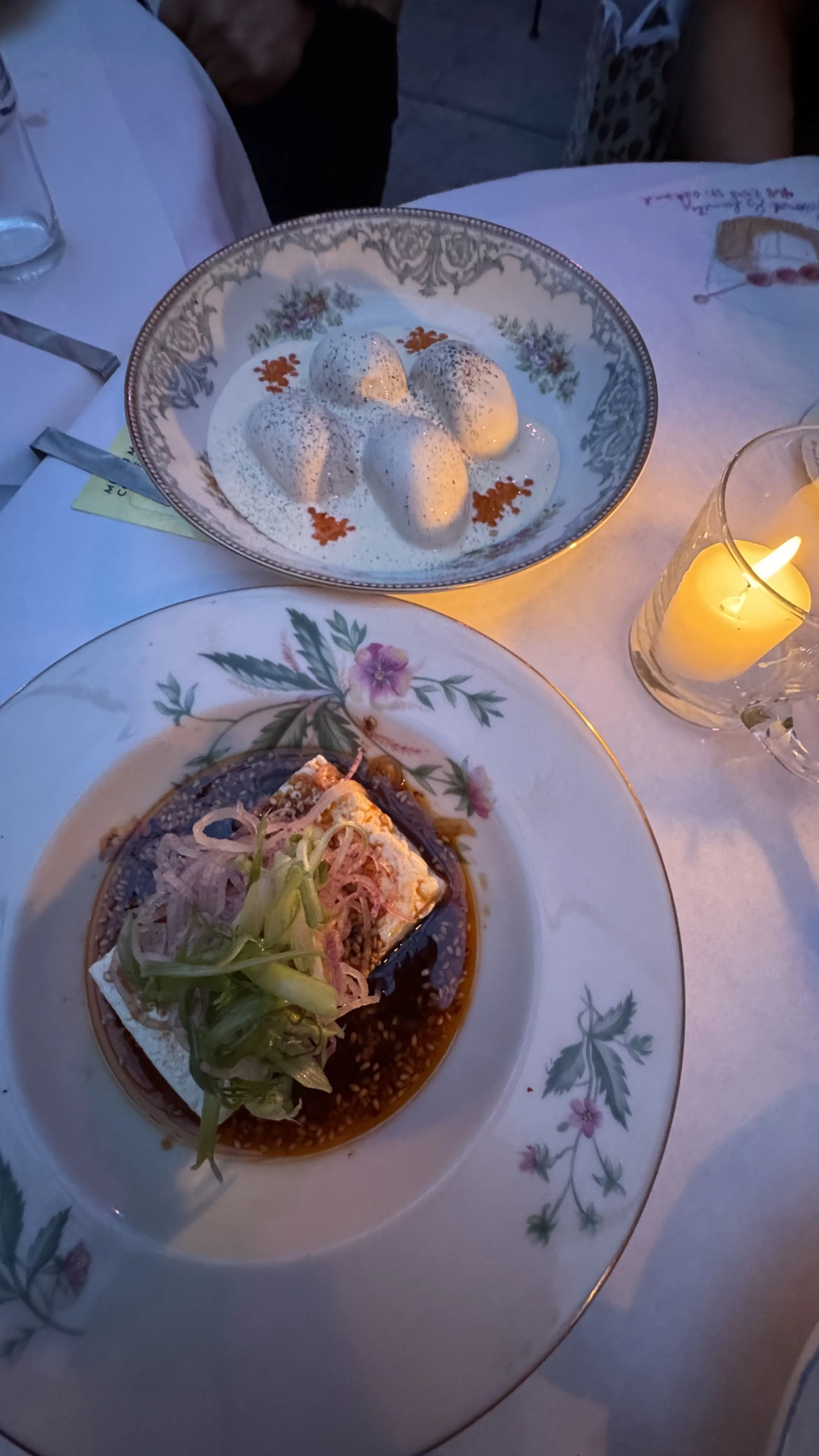 A table set with a plate of dressed fish topped with sliced onions and a soy-based sauce, a bowl of poached eggs with a creamy sauce and red garnishes, and a lit candle in a glass holder, all on a white tablecloth.