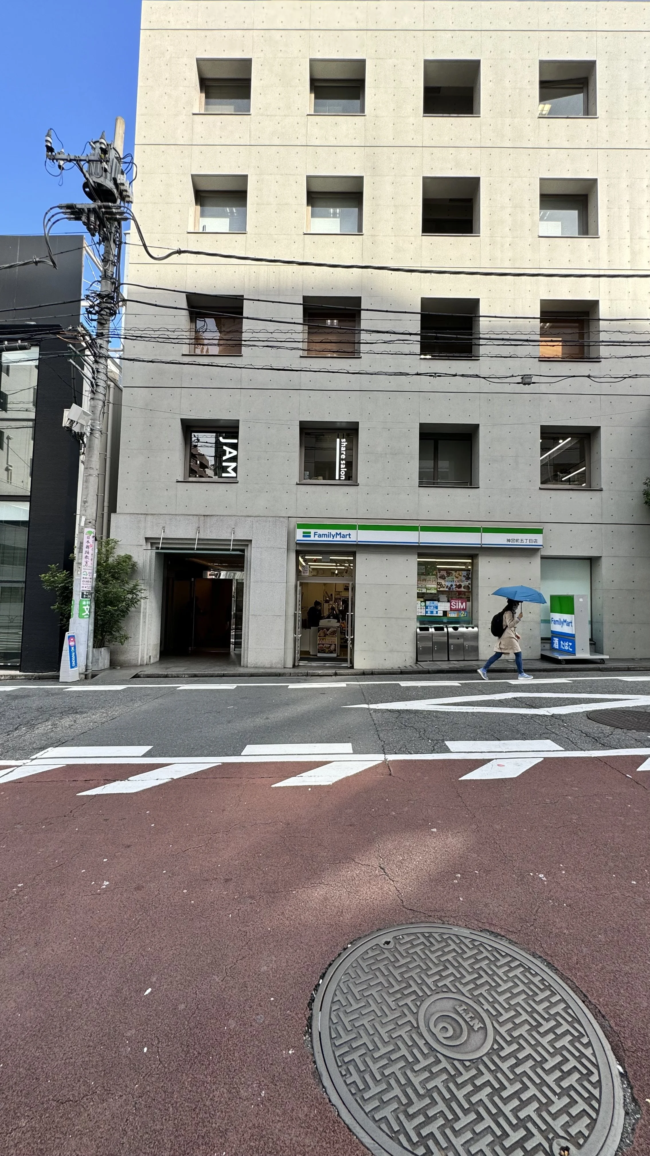 A street view with a woman walking with a blue umbrella in front of a modern building housing a FamilyMart convenience store. The building has multiple small windows and electrical wires are visible overhead.