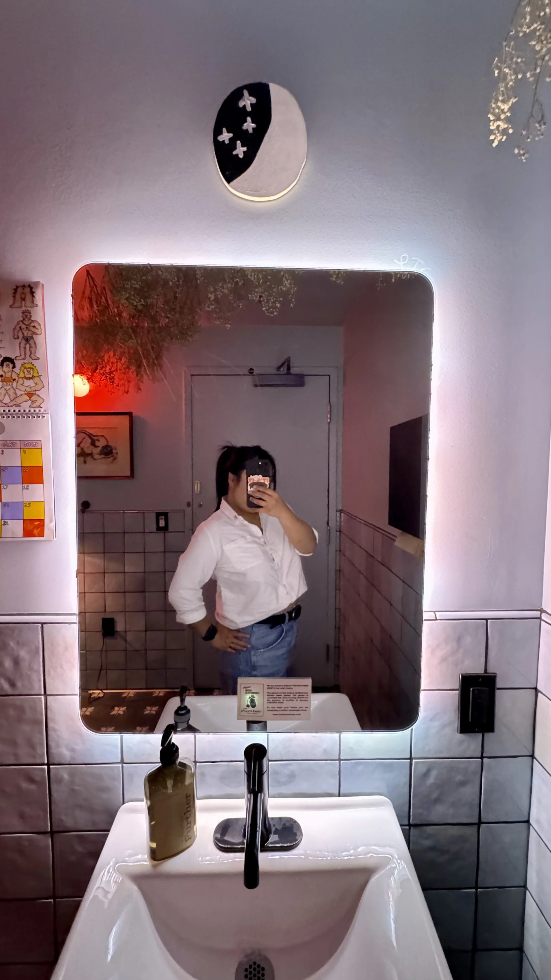 A person taking a mirror selfie in a bathroom, wearing a white shirt and blue jeans. The bathroom has tiled walls, a white sink, and a mirror with LED backlighting. There are decorations on the wall and a red light illuminating part of the room.