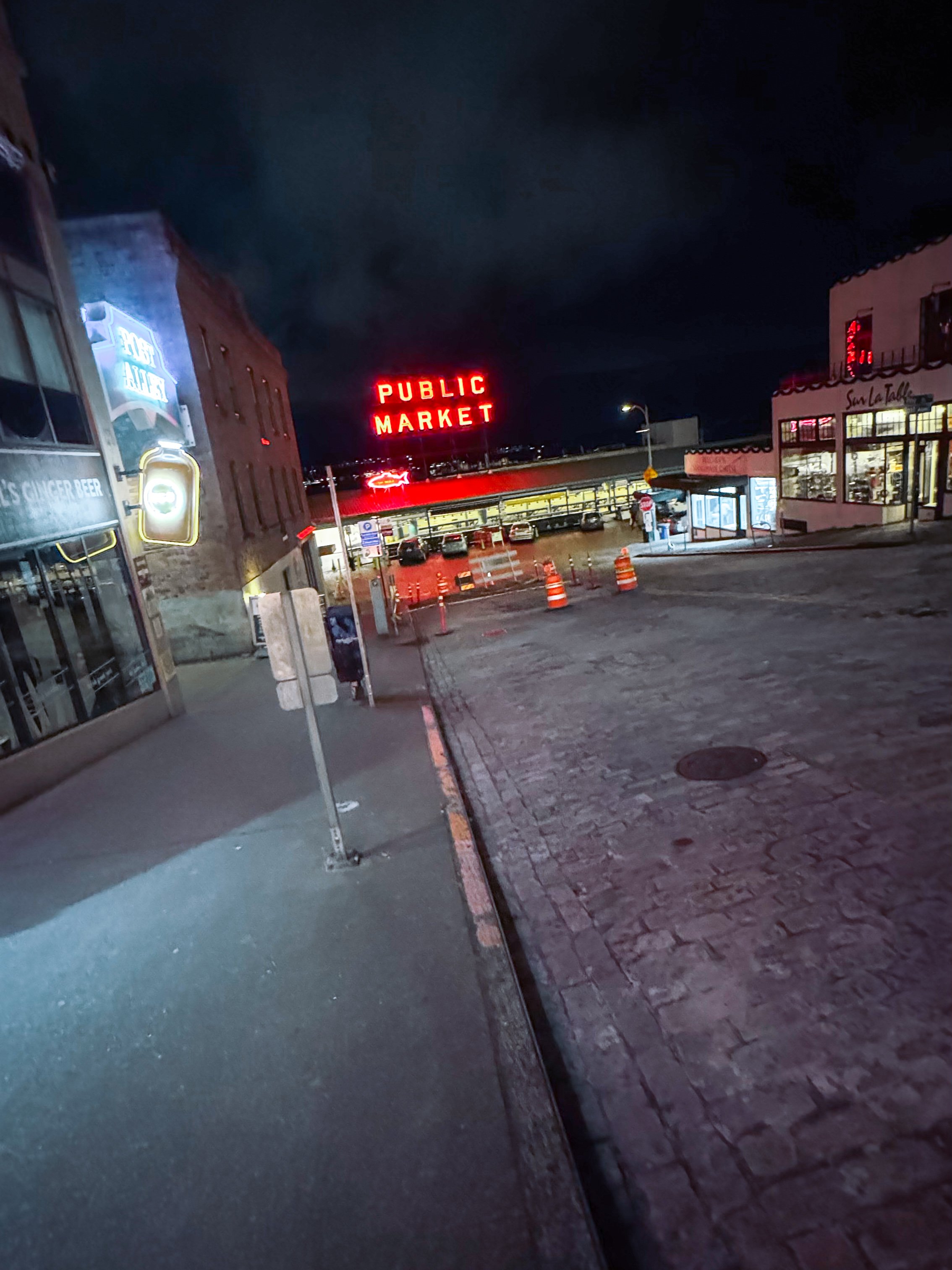 Night scene of a small town street with a large illuminated red sign reading 'Public Market' in the background. The street has some parked cars and orange traffic cones near a parking lot. To the left, there are storefronts with signs, including one with the words 'Post' and 'Surfer Beer,' and a pedestrian standing near a bus stop.