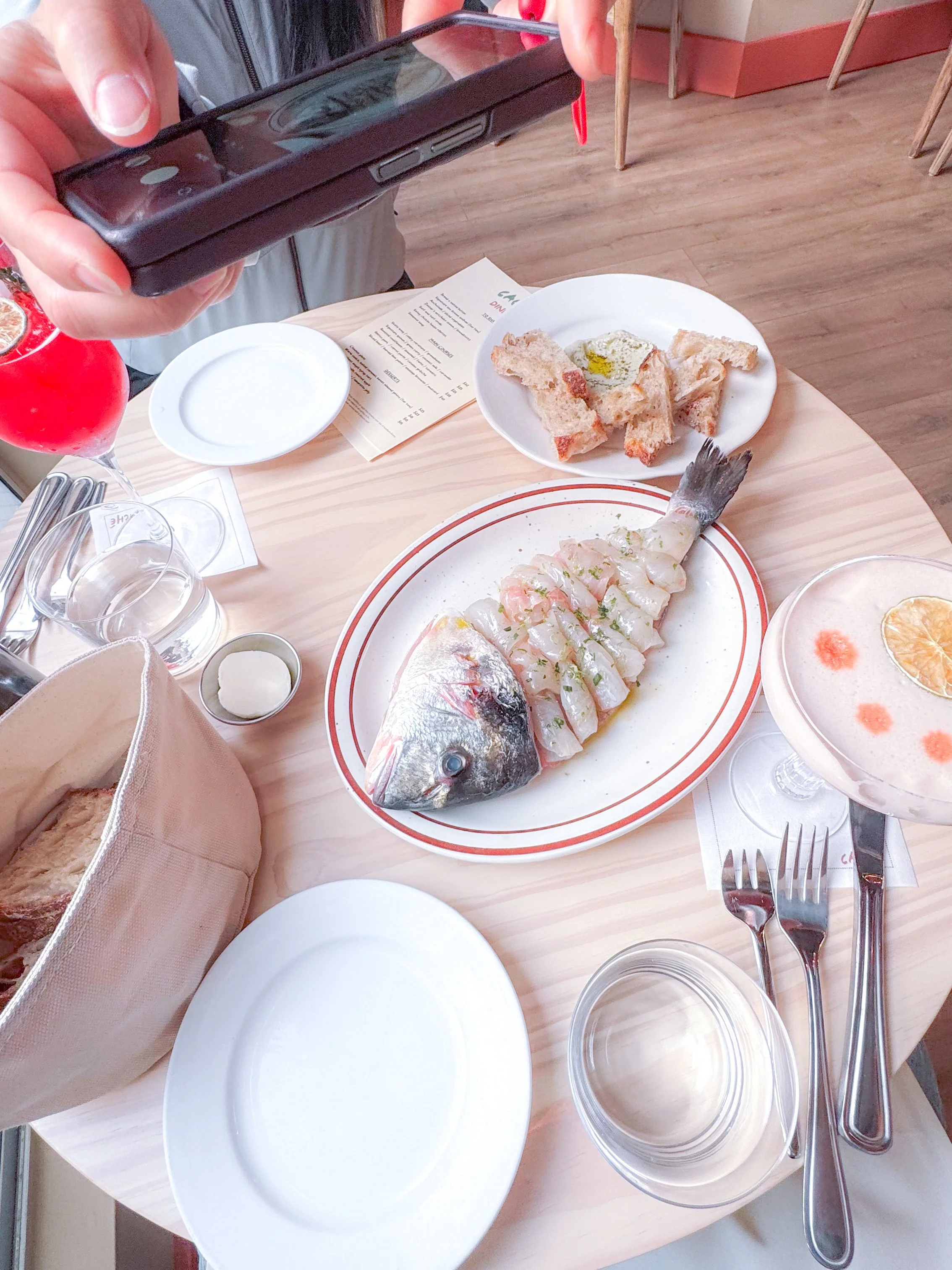 A table with uncooked fish slices on a plate, a plate of bread with eggs, a drink with a slice of lemon, and a person taking a photo with a smartphone.