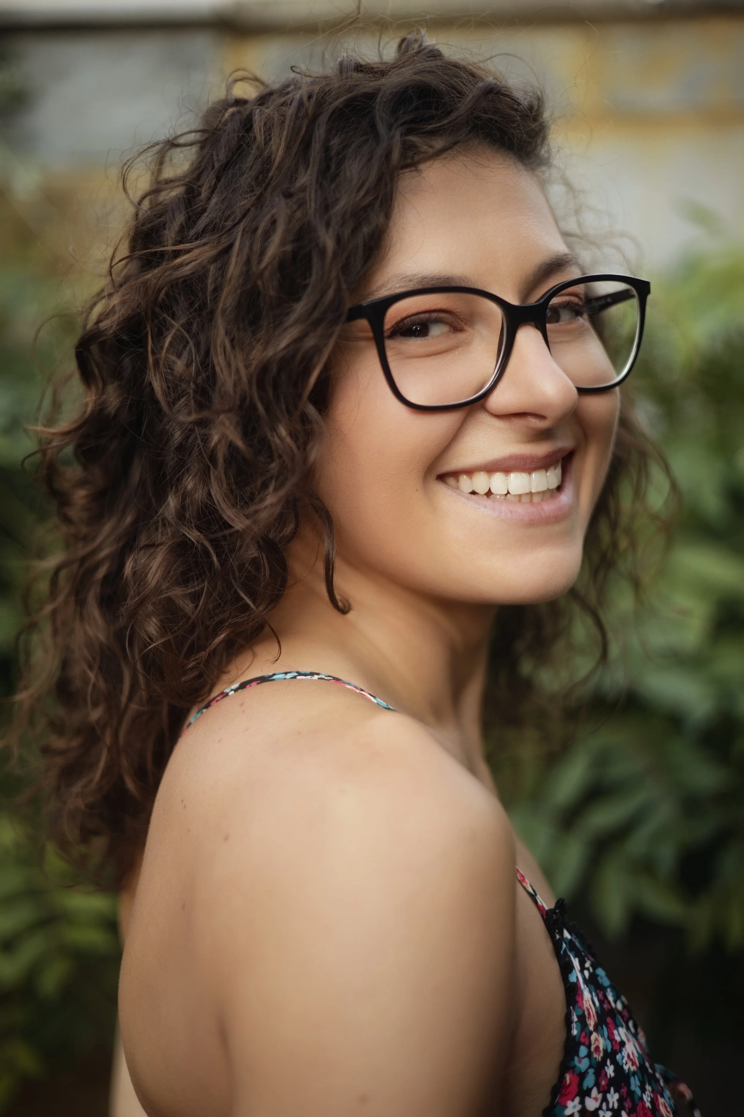 A young woman with curly brown hair and glasses smiling outdoors.