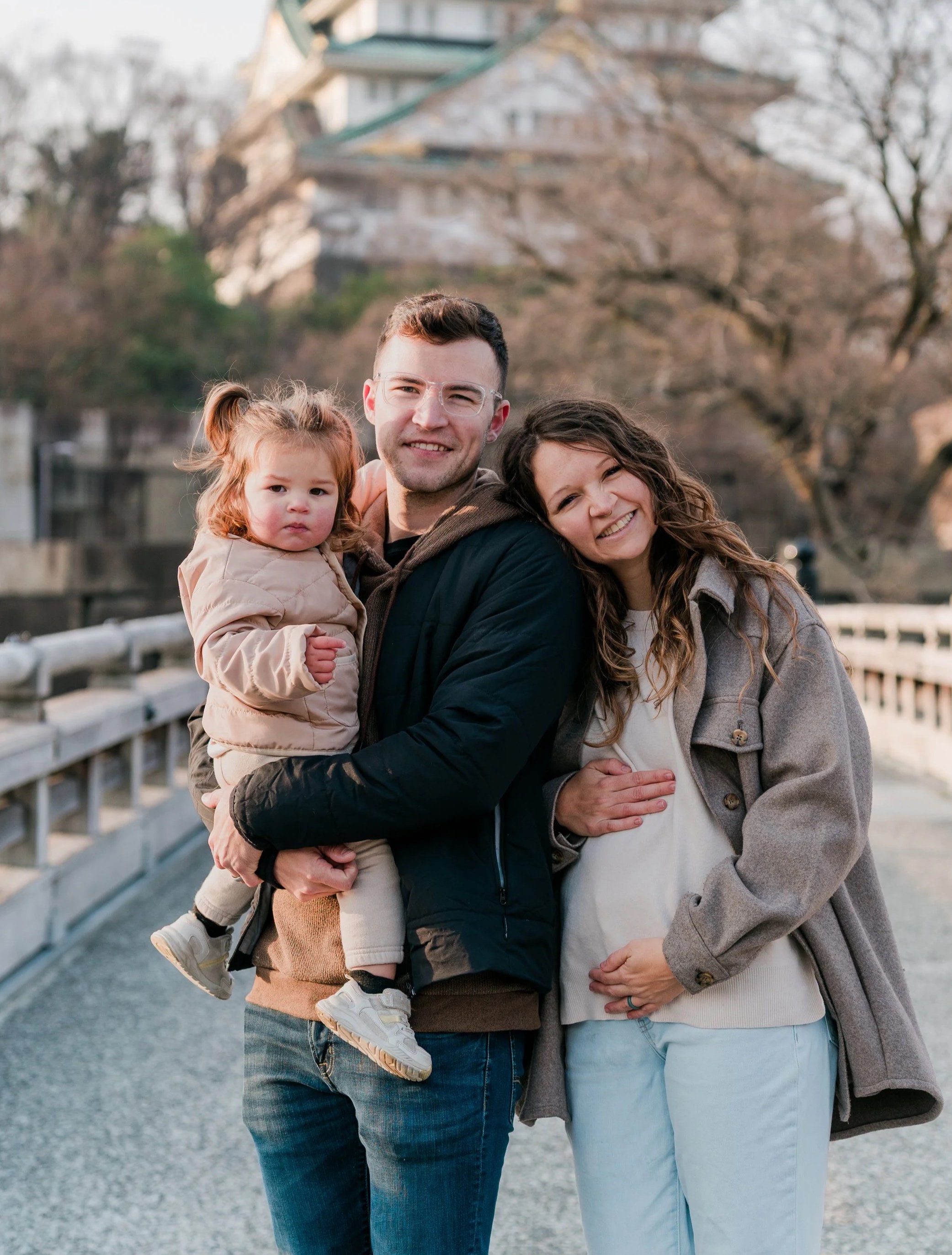 A family of three standing on a bridge outdoors in autumn, smiling at the camera.