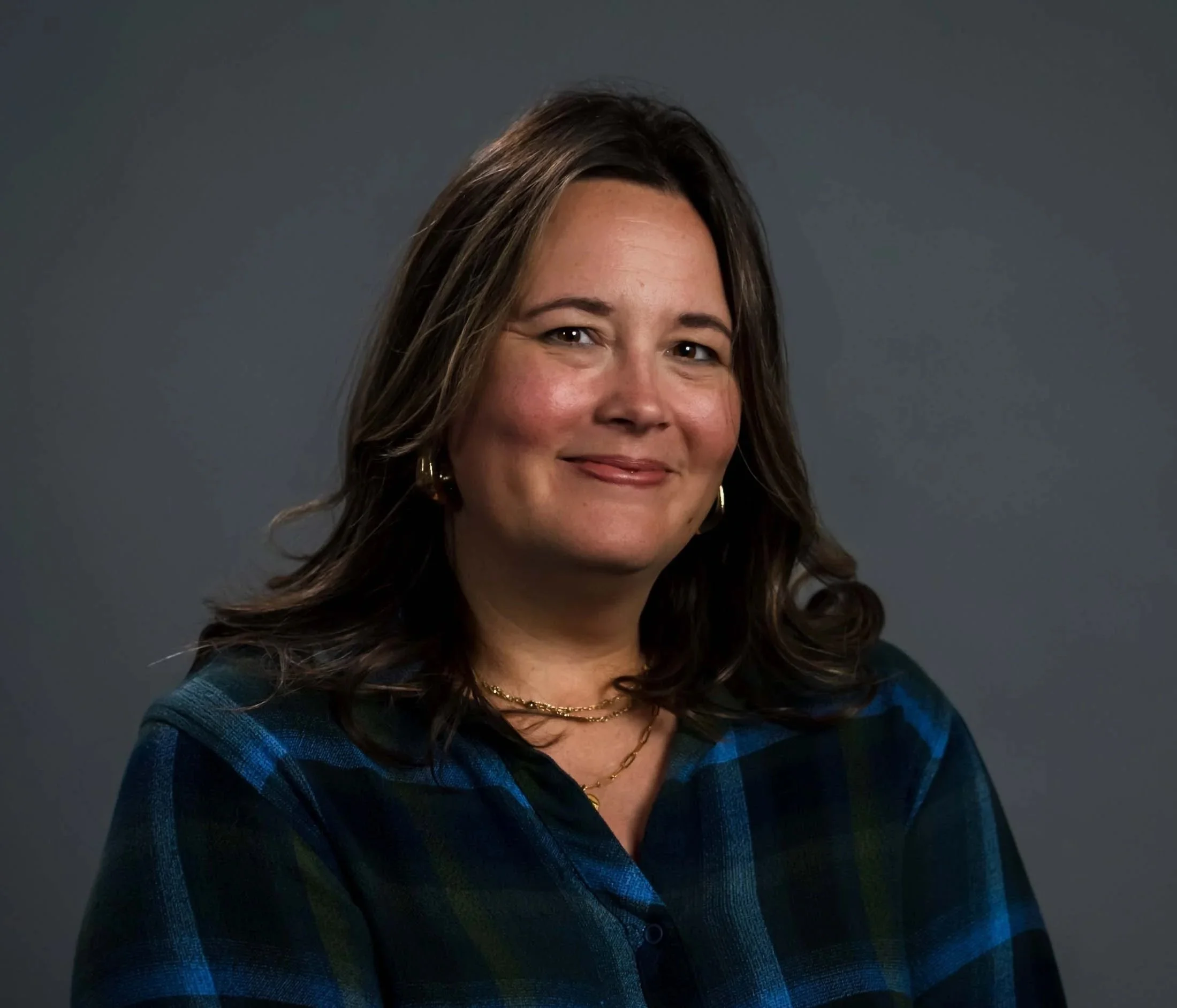 Portrait of a smiling woman with shoulder-length brown hair wearing a blue plaid shirt against a dark gray background.