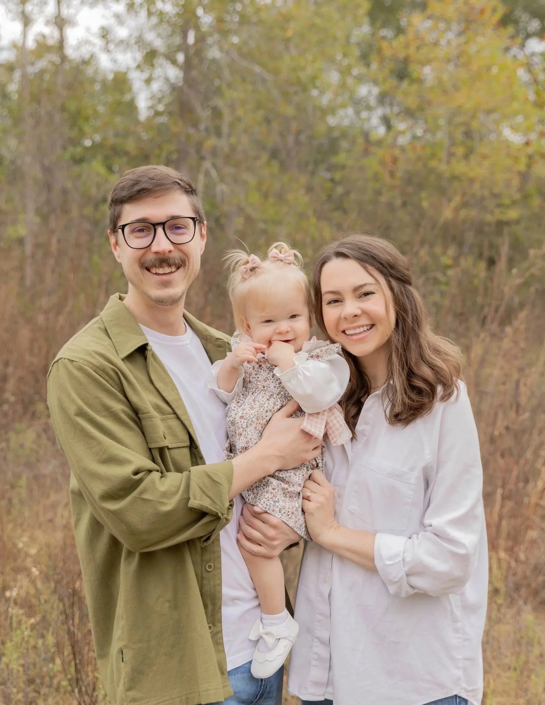A smiling family of three standing outdoors in front of trees, with a man wearing glasses and a woman holding a young girl with blonde hair in pigtails.