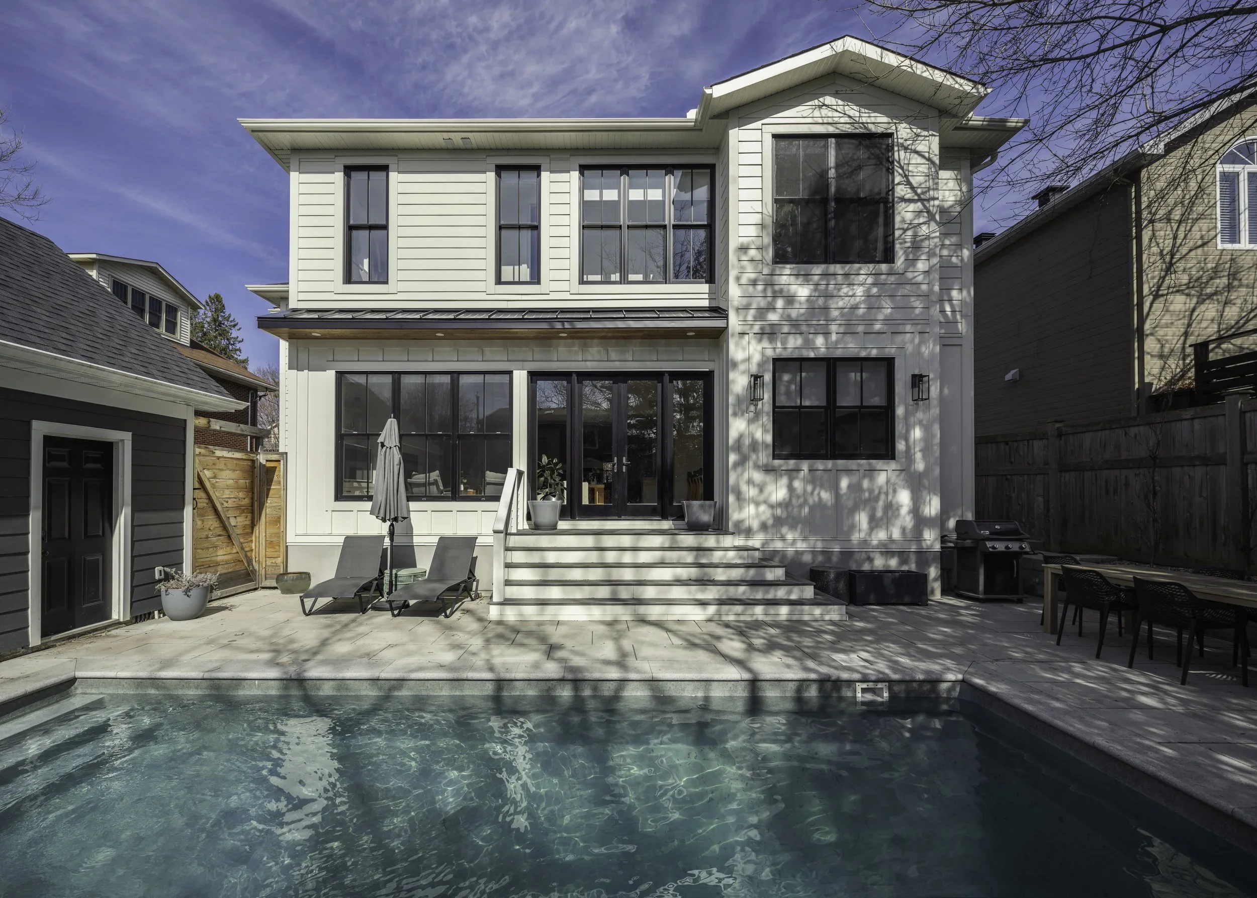 Rear view of a modern, white, three-story house with a backyard pool and outdoor seating area, including lounge chairs, an umbrella, and a dining table with chairs, under a partly cloudy sky.