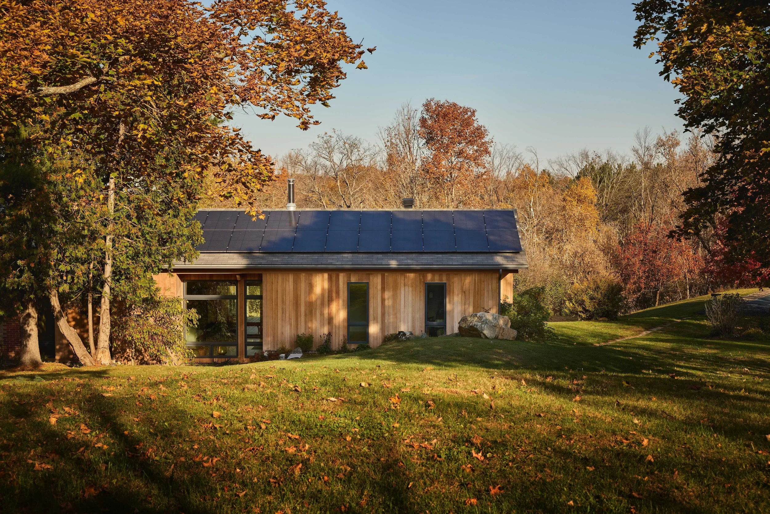 A modern house with a dark roof installed with solar panels, surrounded by trees with autumn foliage, and a grassy lawn in autumn.