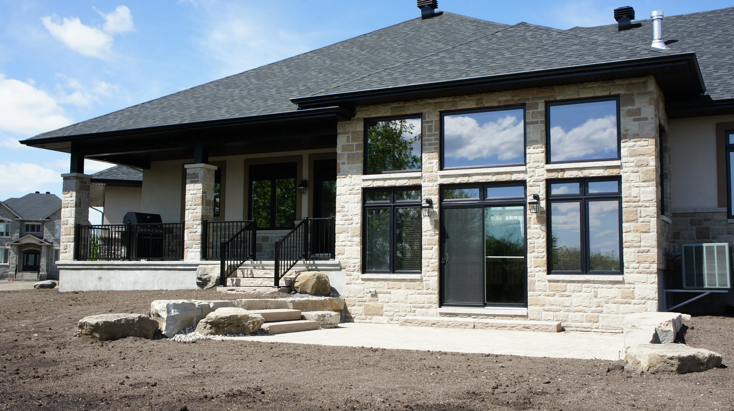 Back view of a new house with large windows, stone accents, a porch with black railing, and a dirt yard with some rocks and steps.