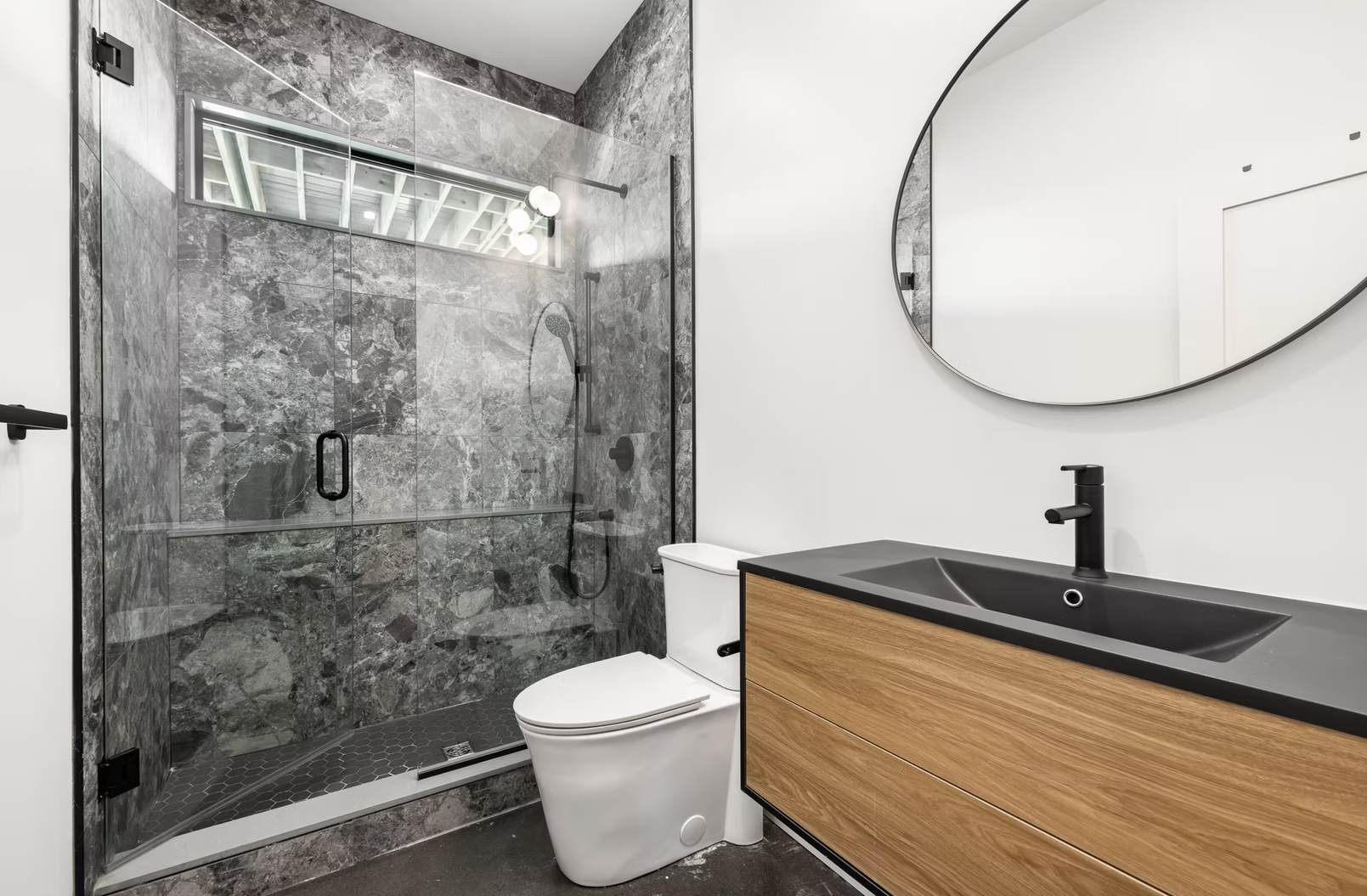 Modern bathroom with a walk-in shower featuring gray marble tiles, a white toilet, a black and wood vanity with a black sink, and a large oval mirror.