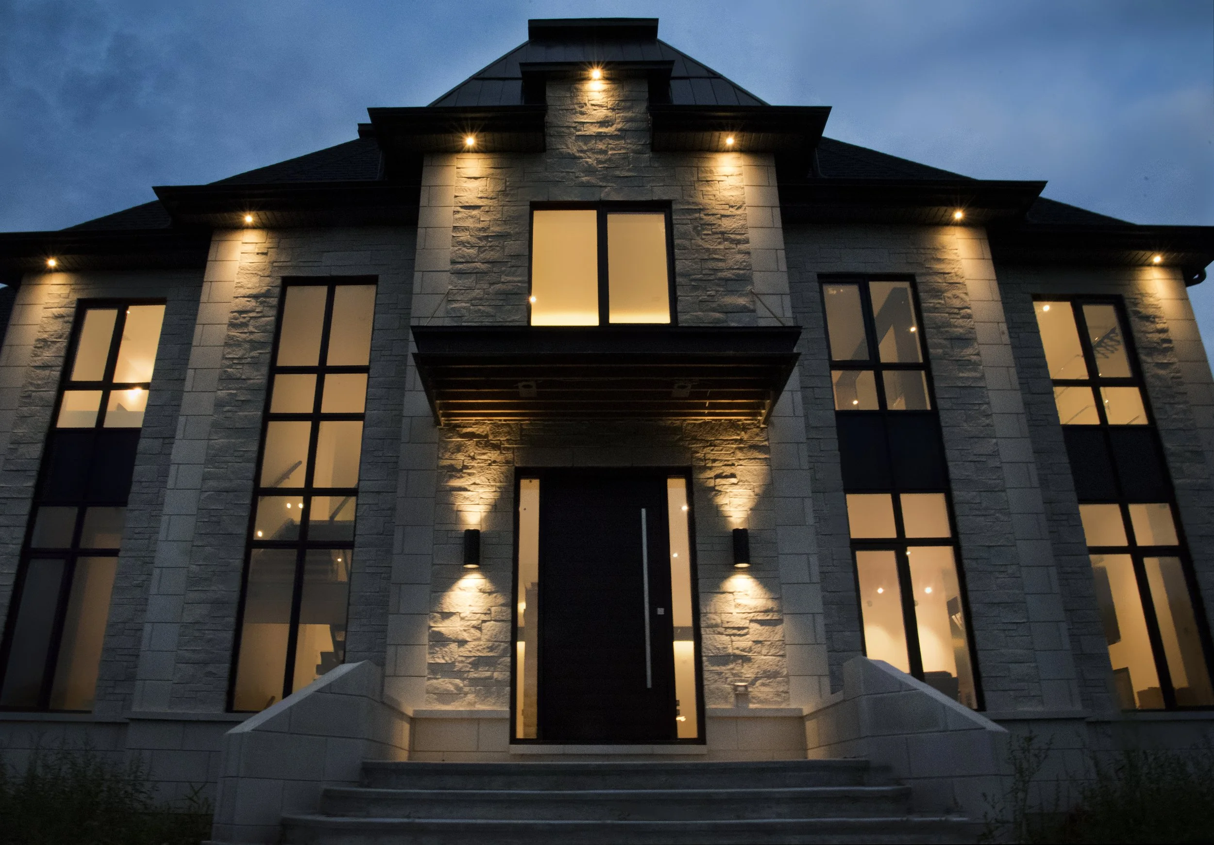A modern multi-story house with large windows illuminated from inside, stone facade, and exterior lighting, during dusk.