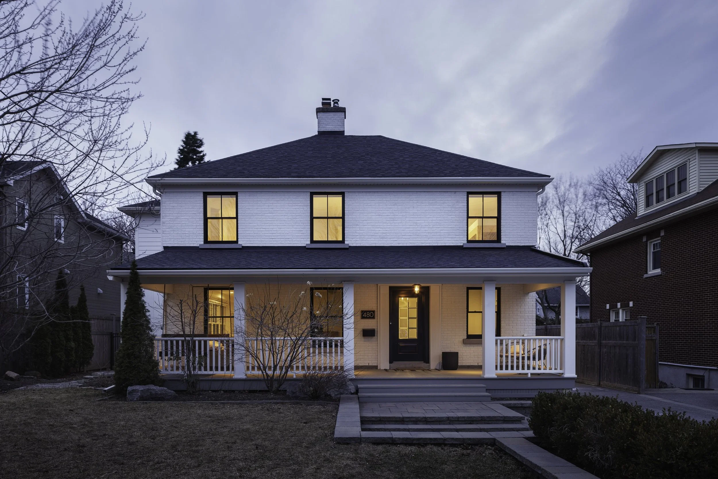 Front view of a two-story white house with a black door and black window frames, illuminated from inside, during dusk. The house has a porch with white railing, a path leading to the steps, and a yard with some bushes and small trees.