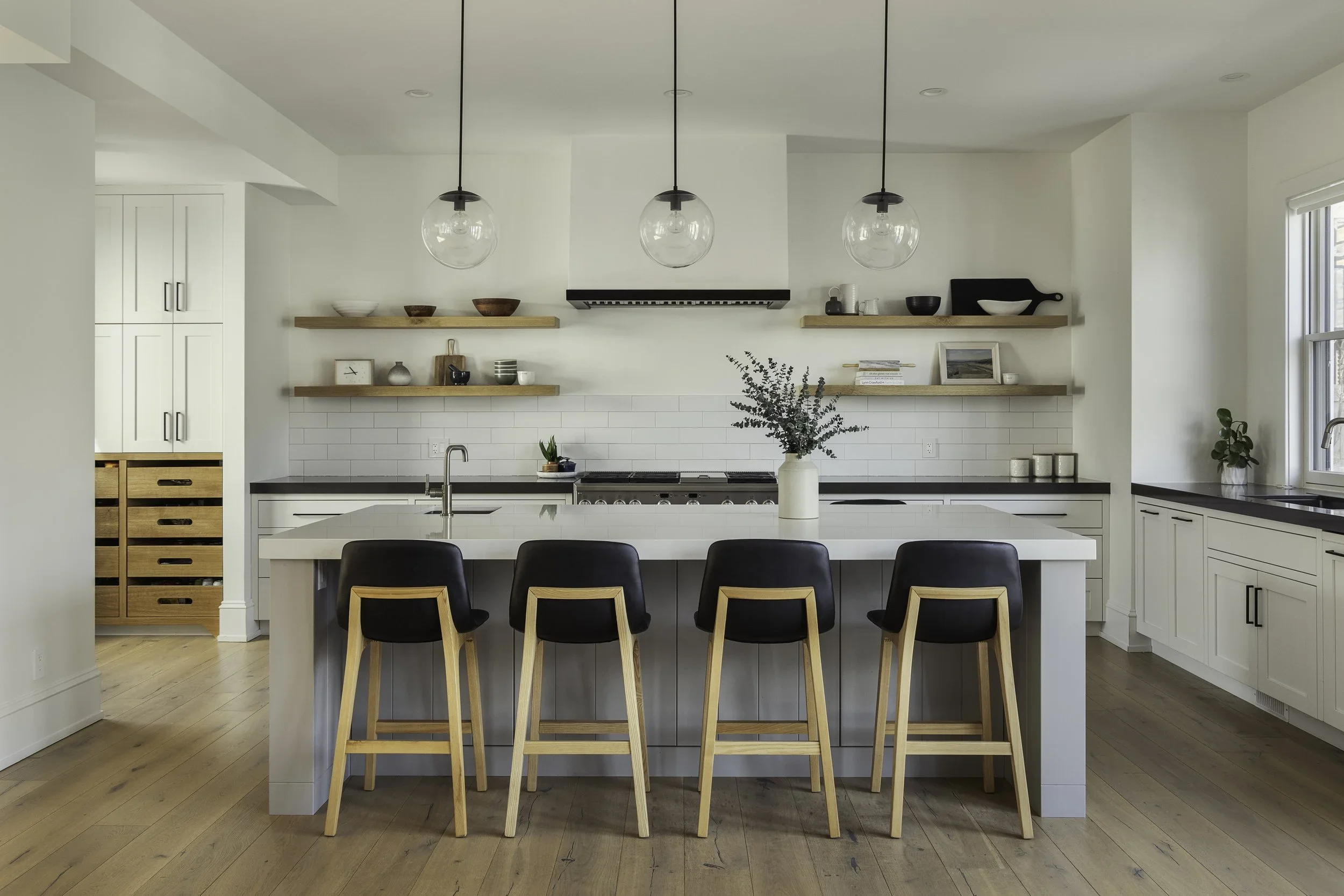 Modern kitchen with white cabinets, black countertops, open wooden shelves, a large island with black and wooden bar stools, pendant lights, a window, and minimal decoration.