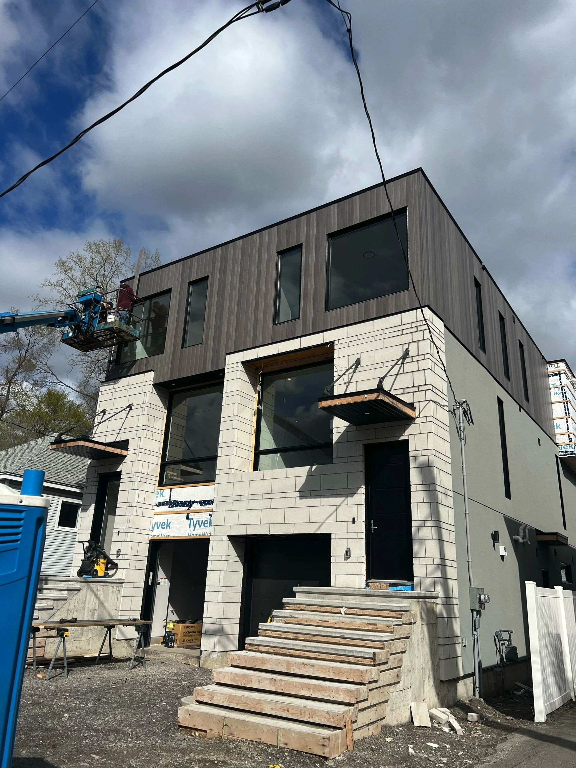 Under construction modern multi-story house with white brick exterior, large windows, and wooden accents, with construction tools and materials around, and worker on lift