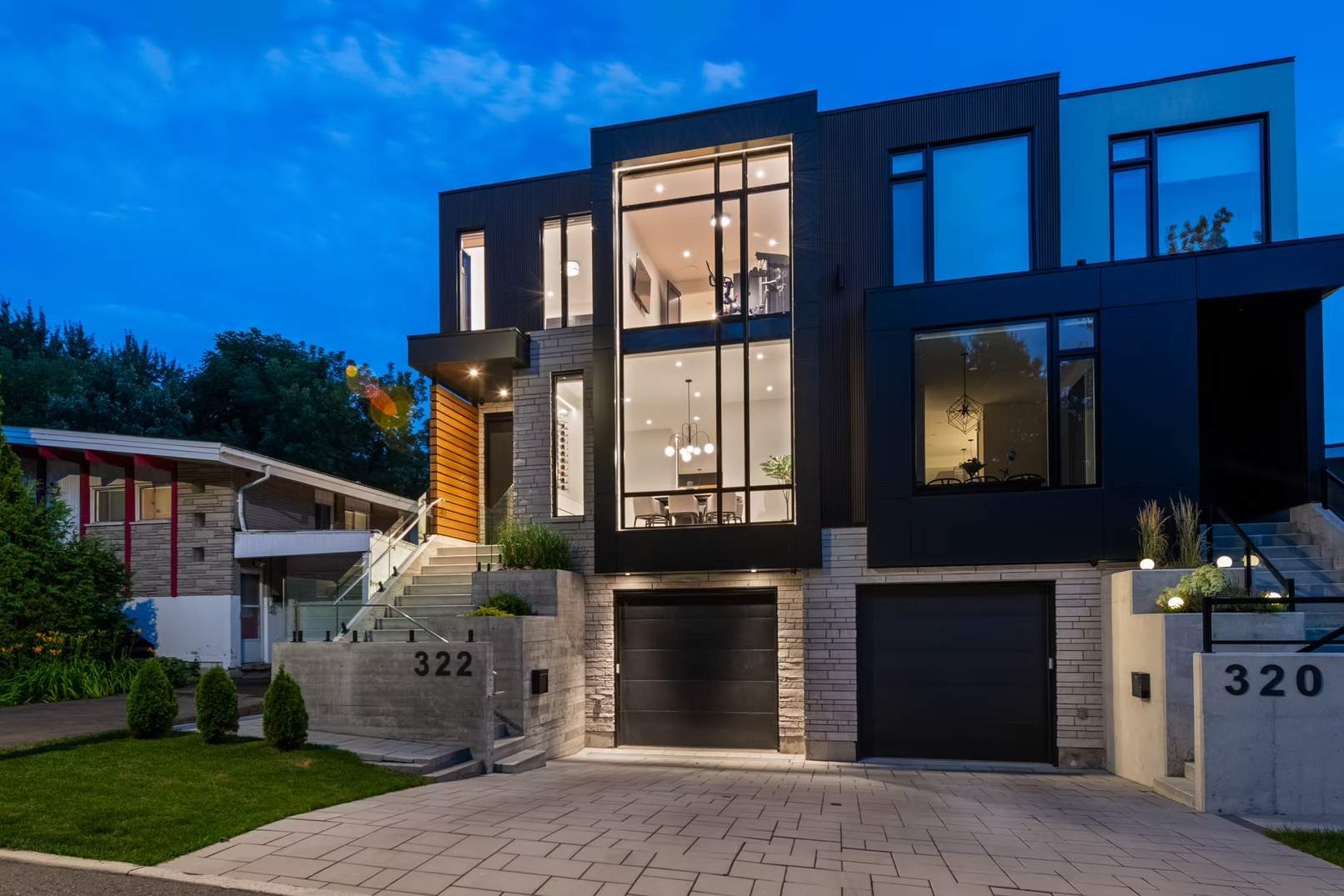 Modern multi-story house with large glass windows, illuminated interior, and landscaped front yard at dusk.