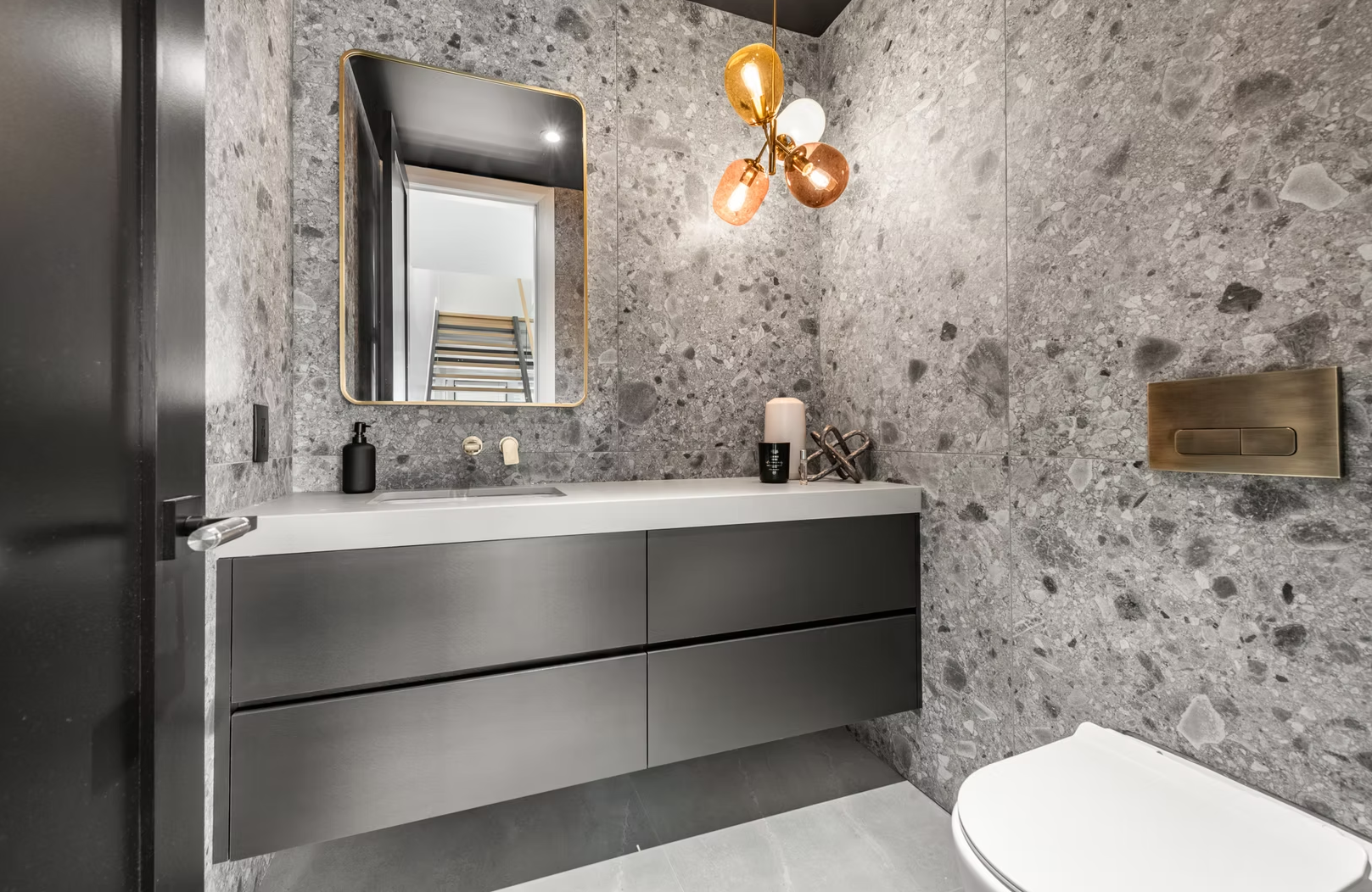 Modern bathroom with gray textured wall and floor, wall-mounted gray vanity with a white countertop, black soap dispenser, and decorative items, mirror with gold trim, unique multi-colored glass pendant light, and a partially visible toilet
