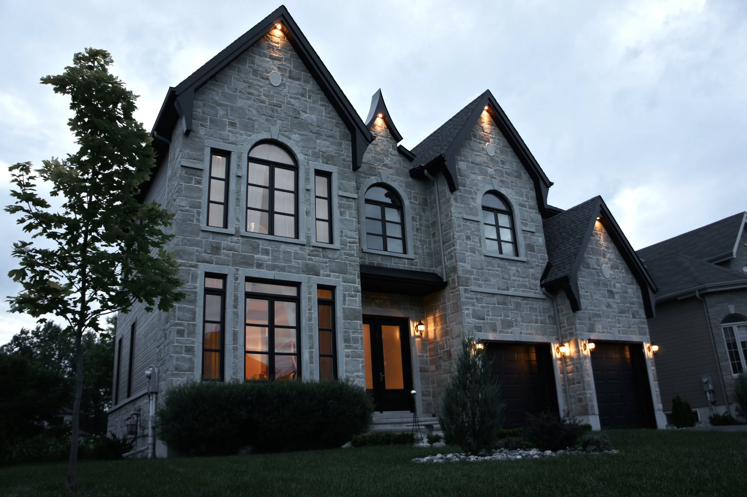 A two-story stone house with multiple lit exterior lights at dusk, large arched and rectangular windows, and a well-maintained lawn with shrubs and a young tree in the front.