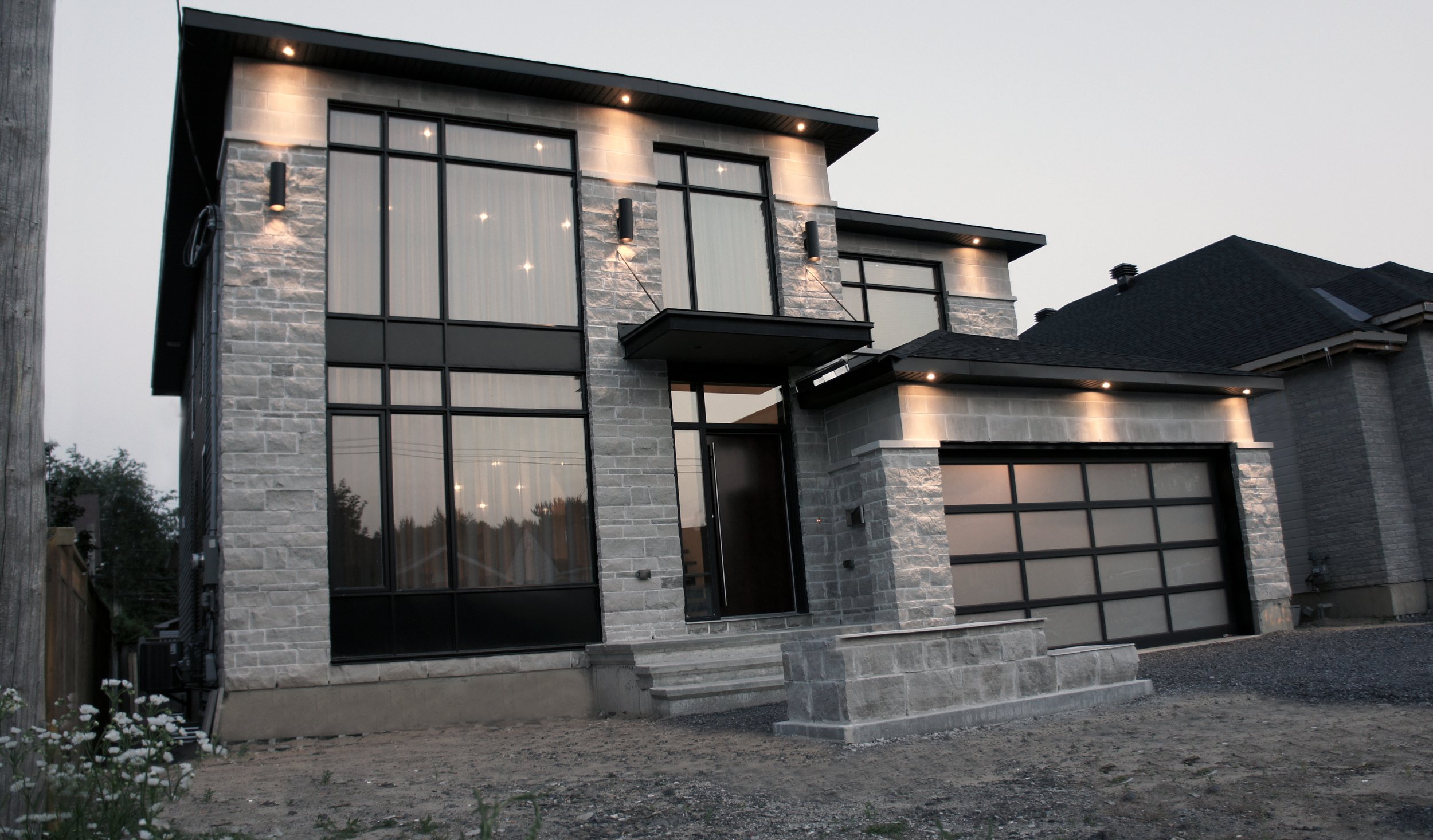 A modern, two-story house with a stone facade, large black-framed windows, and a garage with a black paneled door. Exterior lights illuminate the house, and the front yard is unfinished with dirt and small plants.