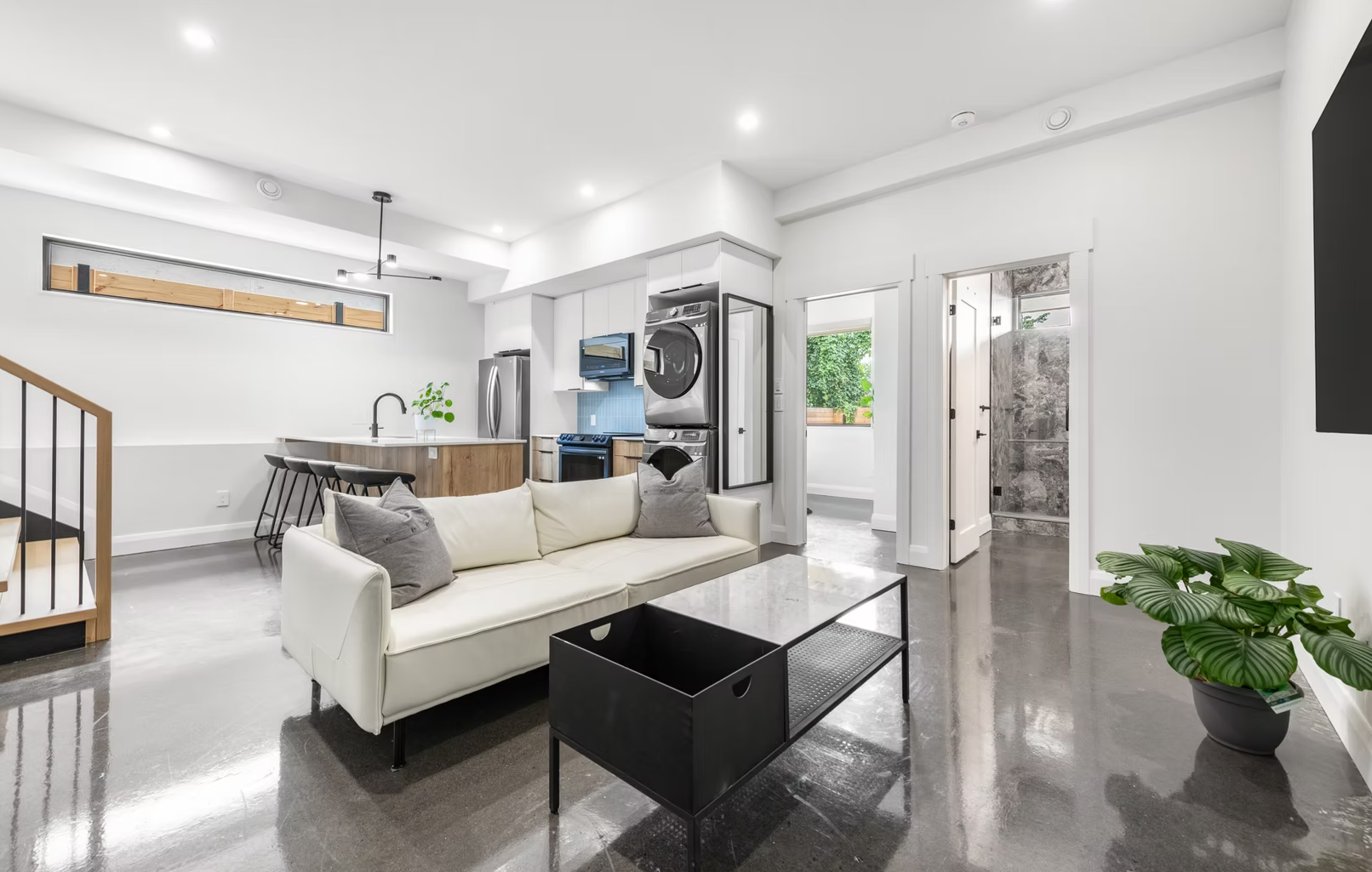 Modern open-concept living room and kitchen with white walls, polished concrete floors, a white sofa with gray pillows, black coffee table, potted plant, kitchen with wooden accents, stainless steel appliances, and laundry stacked near the kitchen.