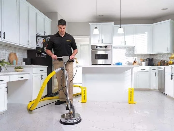 A man cleaning a kitchen floor with a Stanley Steemer carpet cleaner. The kitchen has white cabinets, a central island, and stainless steel appliances.