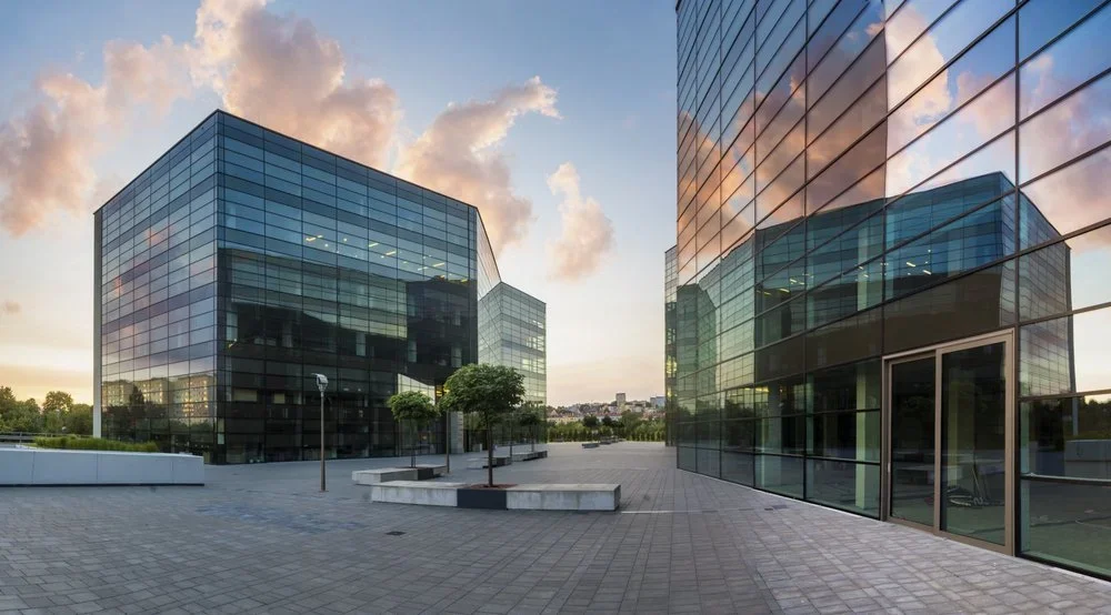 Modern glass office buildings reflecting the sky at sunset, with a paved plaza area and trees in the foreground.