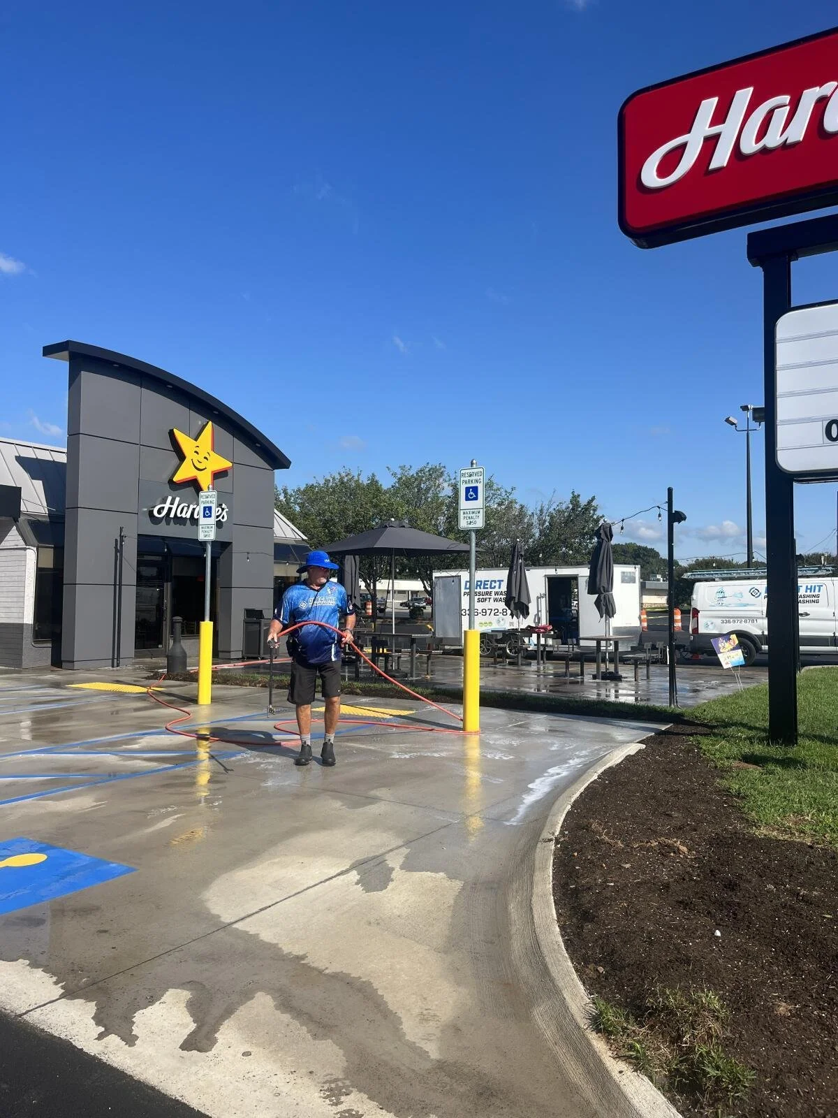 A man in a blue shirt, black shorts, and a blue hat is cleaning the parking lot with a hose outside a Hardee's restaurant on a sunny day. The restaurant has a yellow star with a smiley face logo. There are parking spots, a reserved parking sign for handicapped, and tables with umbrellas in the background.
