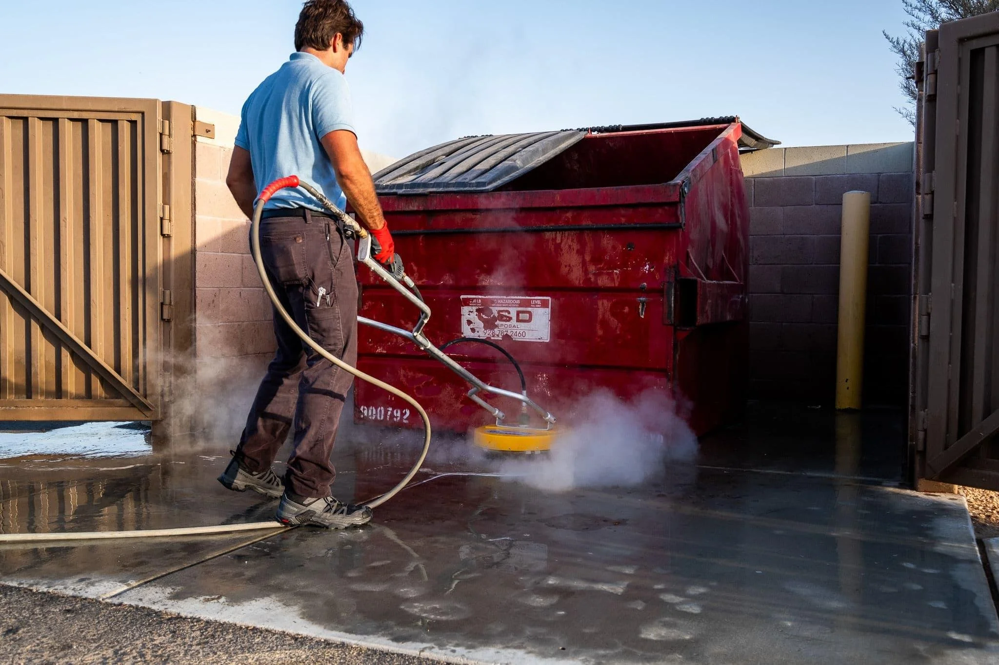 A man wearing a blue shirt, brown pants, and red gloves is pressure washing a stained concrete driveway near a red dumpster in a residential backyard, with a wooden gate on the left and a cinder block wall on the right.