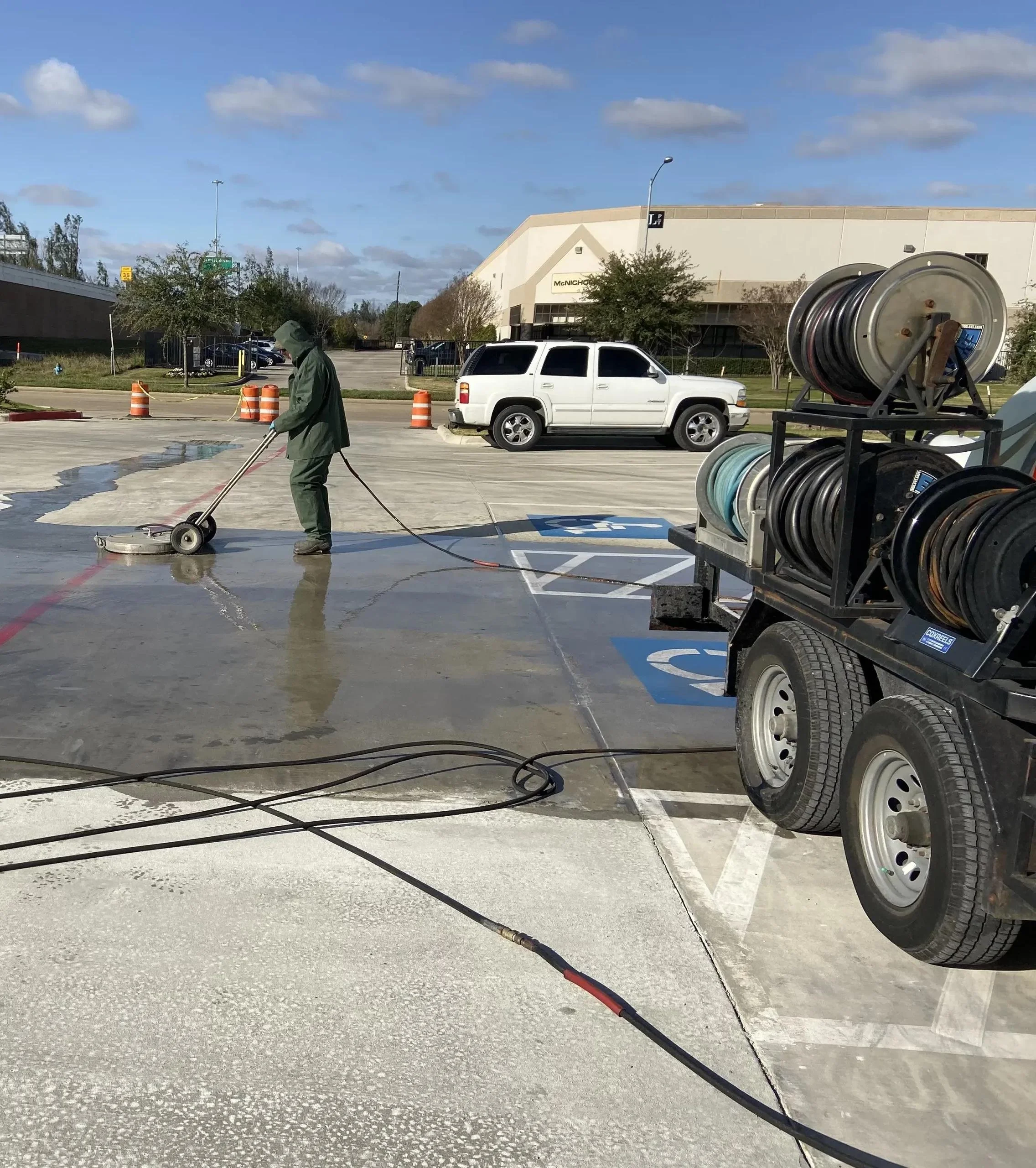 A worker in a green jacket and hood pressure-washing a parking lot. The scene includes parked cars, a building in the background, orange traffic cones, and a cart with multiple hoses.
