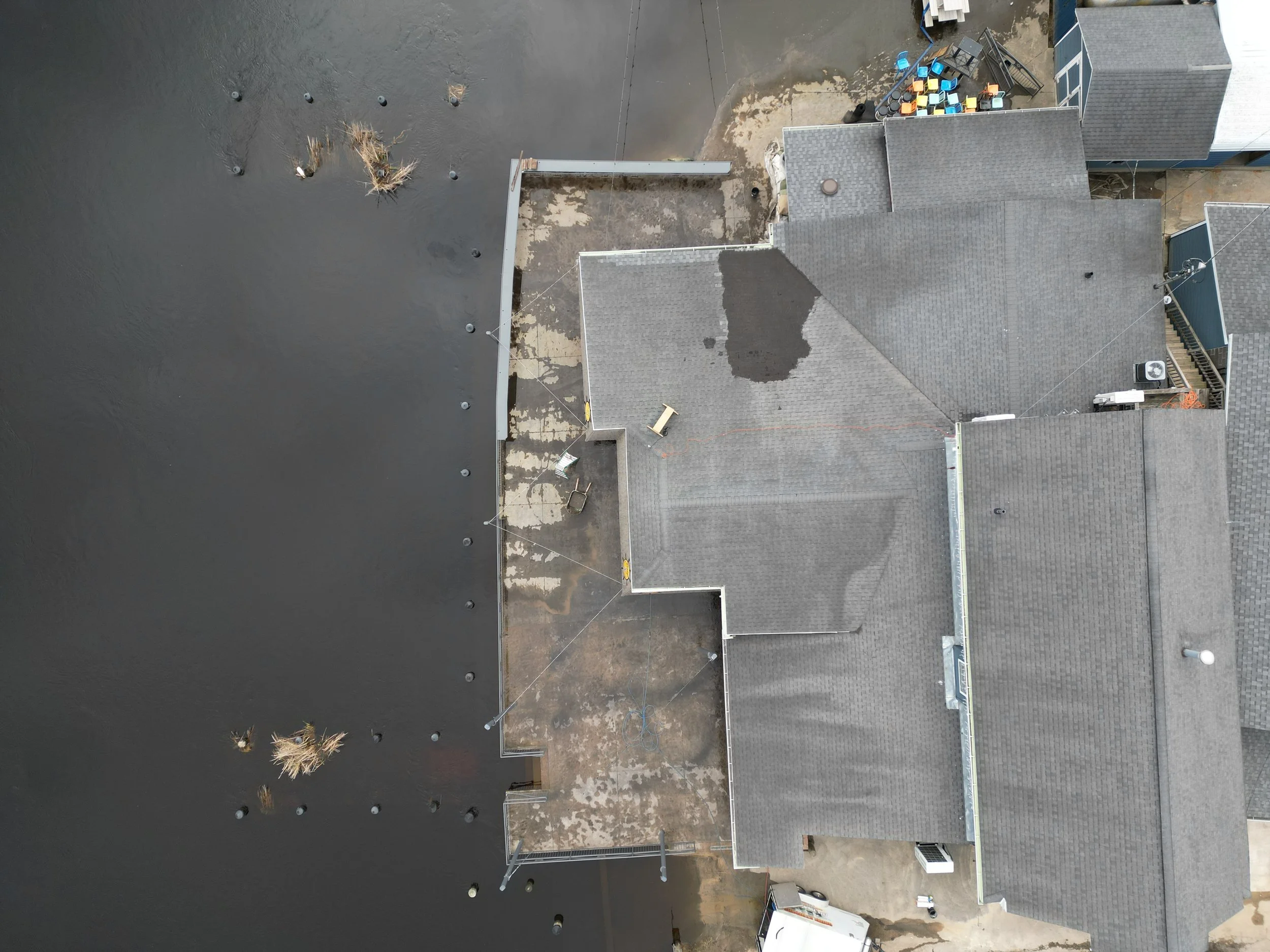An aerial view of rooftops with grey shingles, some with trees, satellite dishes, and various equipment, alongside a large black surface possibly a water body or asphalt.