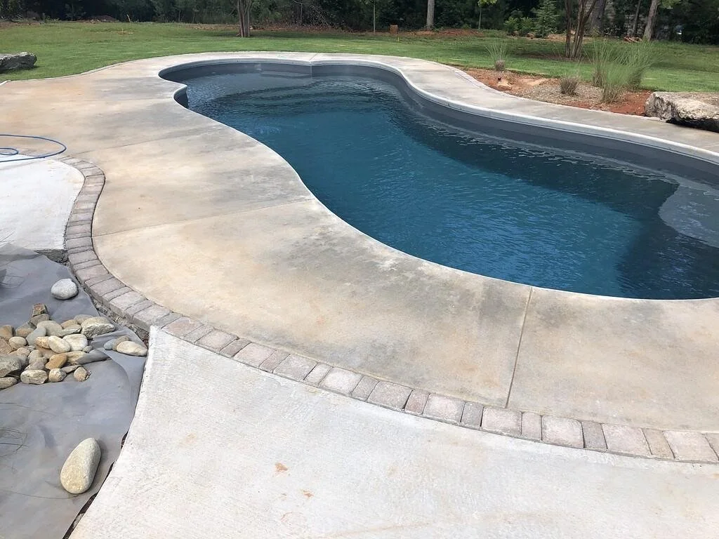 A backyard swimming pool with a concrete deck and landscaping around it, including rocks and grass.