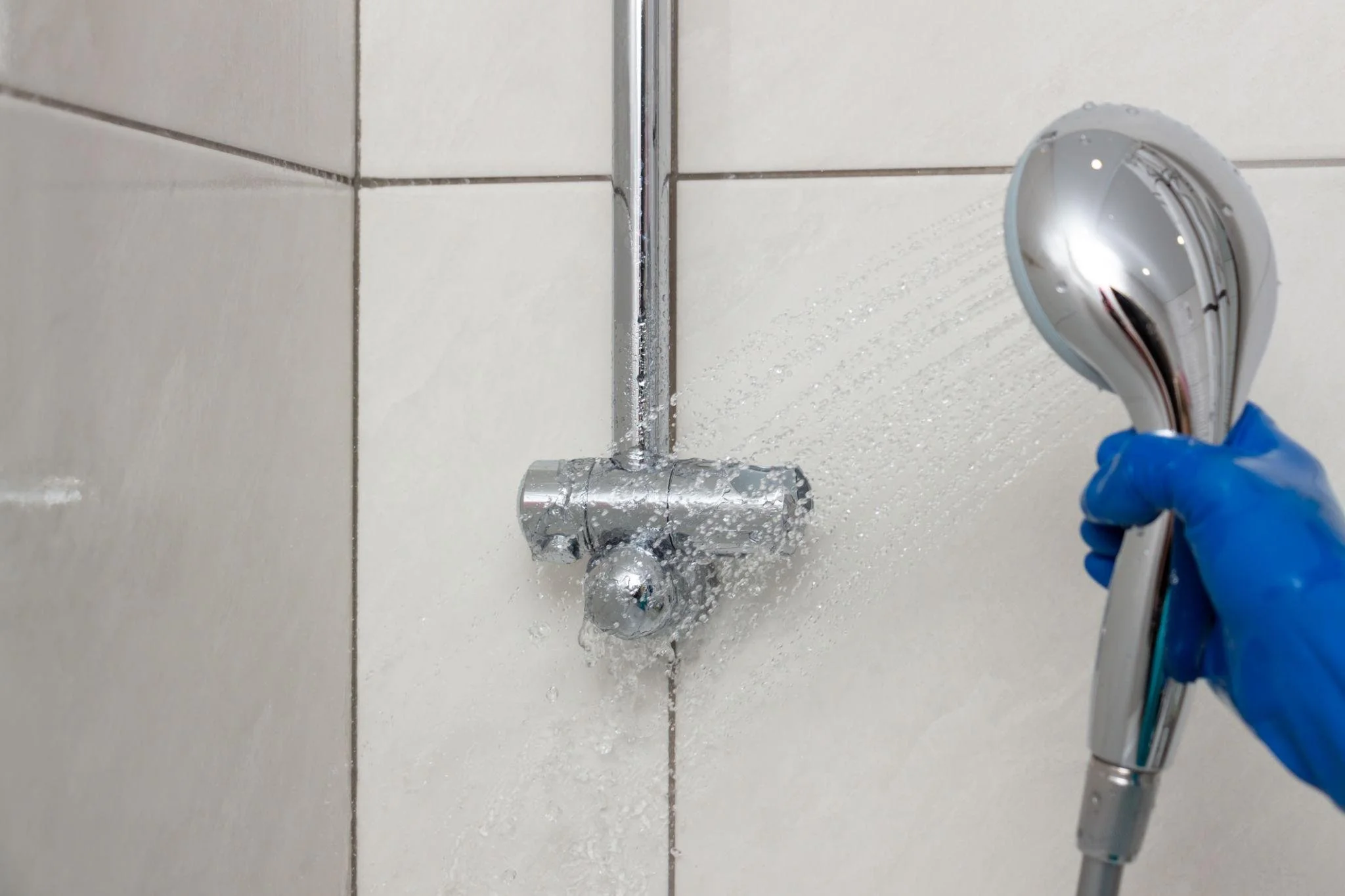 A person wearing blue gloves holding a showerhead in a bathroom with white tiled walls, water spraying from the showerhead.