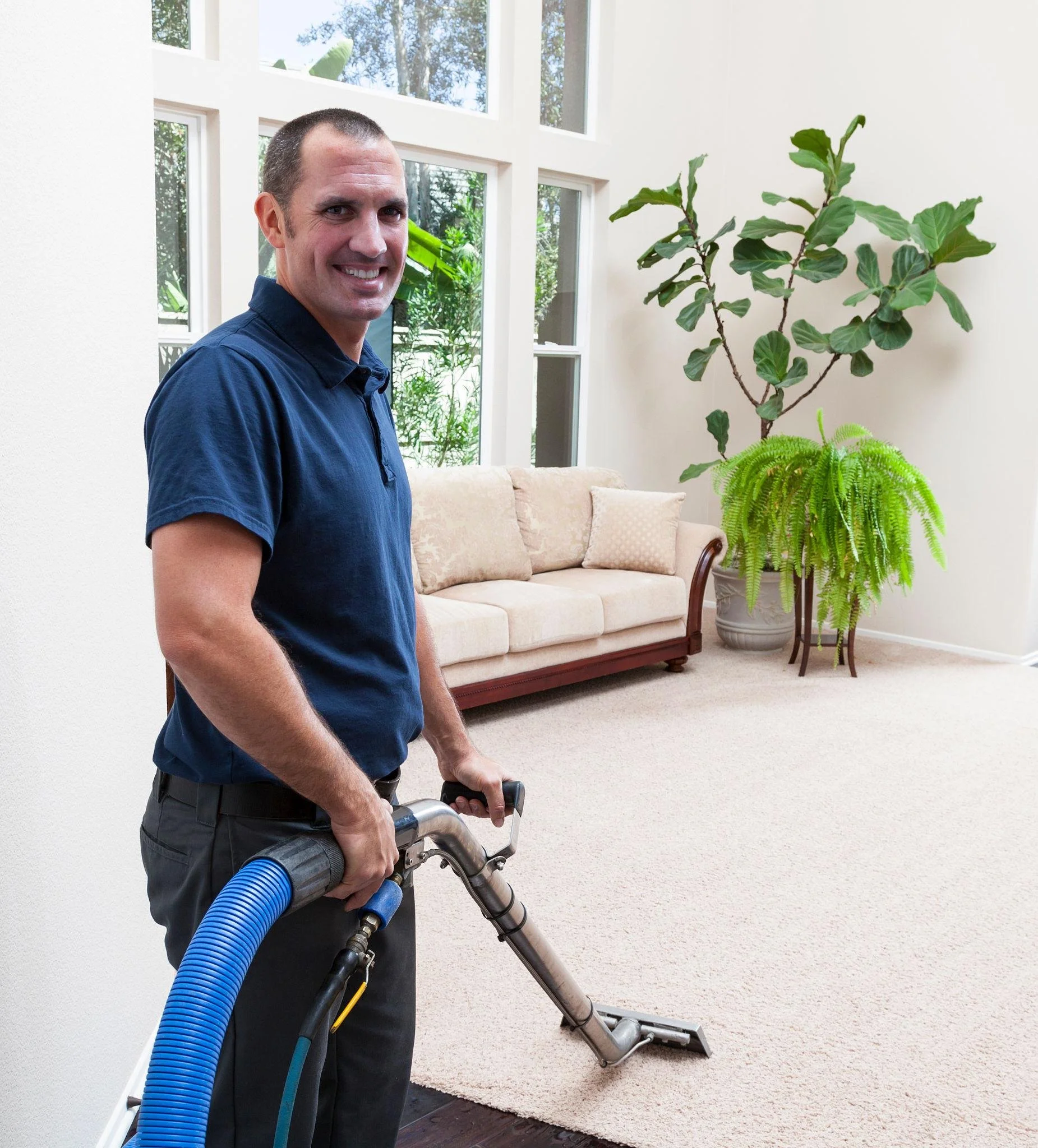 A man smiling while holding a vacuum cleaner in a living room with a beige sofa, large window, and green plants.