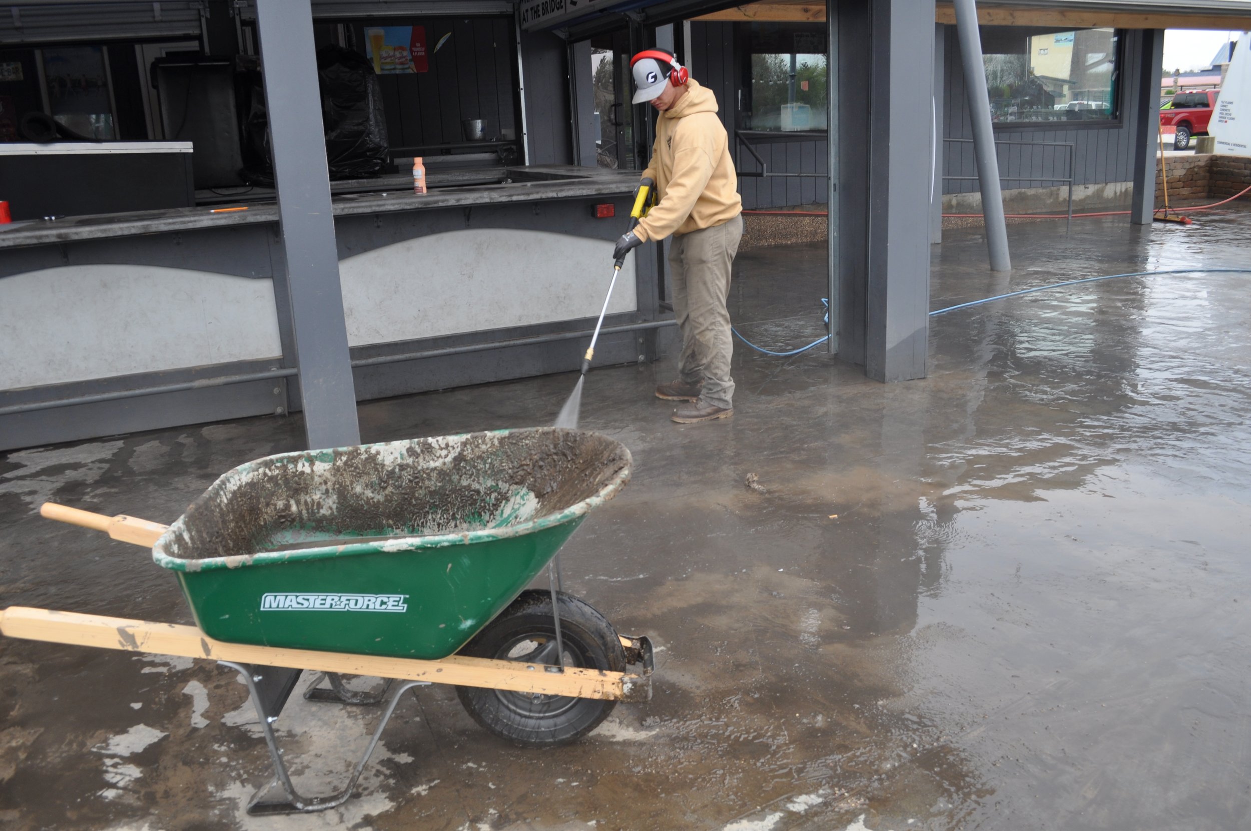 A person pouring concrete on a construction site, standing on wet concrete, wearing a beige hoodie, gray pants, gloves, a helmet, and headphones. There is a wheelbarrow and other construction tools around.