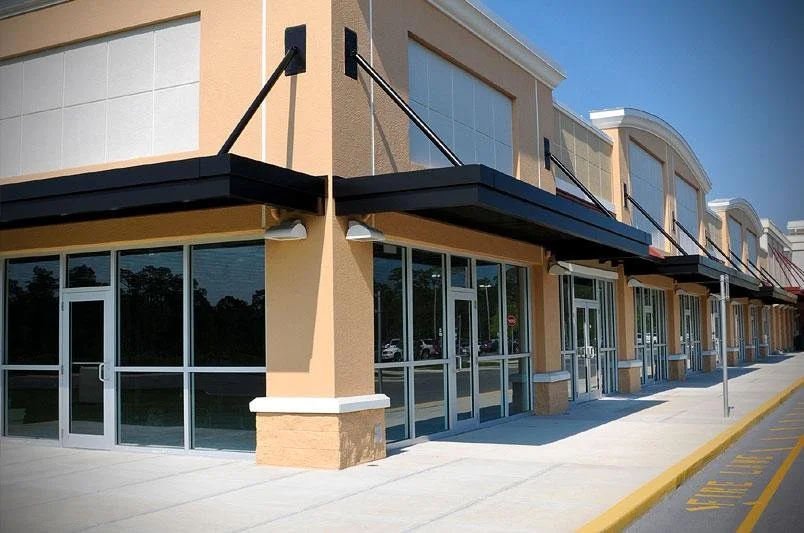 Commercial storefront with glass windows, doors, black awnings, and a sidewalk, in a shopping plaza.