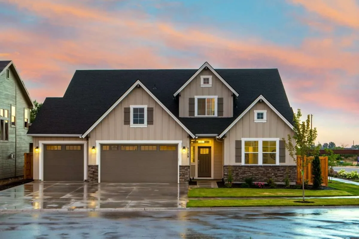 A modern suburban house with a three-car garage, beige siding, dark roof, and a well-maintained lawn, during sunset with colorful sky.
