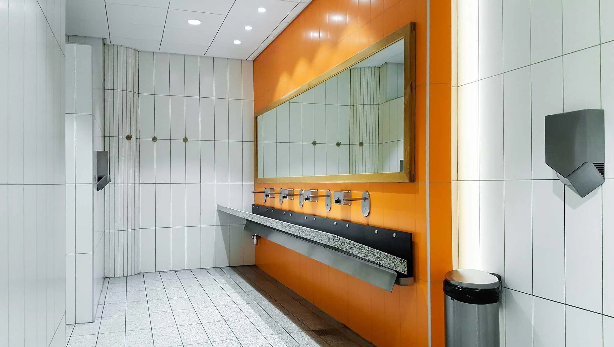 Public restroom with white tiled walls and floor, orange accent wall with a large mirror, and a black countertop with sinks and multiple paper towel dispensers.