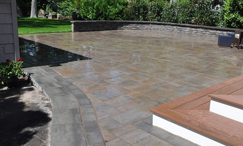 A backyard patio with wet stone tiles, a curved concrete border, a small flower bed with red flowers, some stairs with brown wood steps, a gray chair, and greenery including trees and bushes in the background.