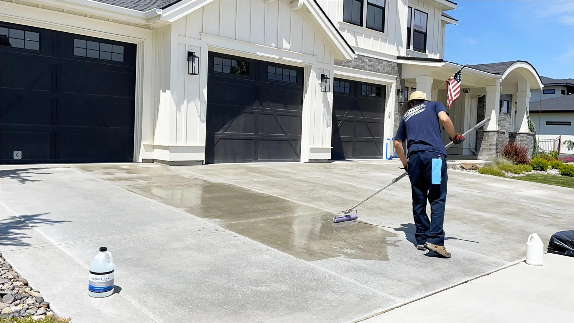 A worker applying a coating or sealing agent to a concrete driveway in front of a modern house with a black garage door and an American flag, under clear blue skies.