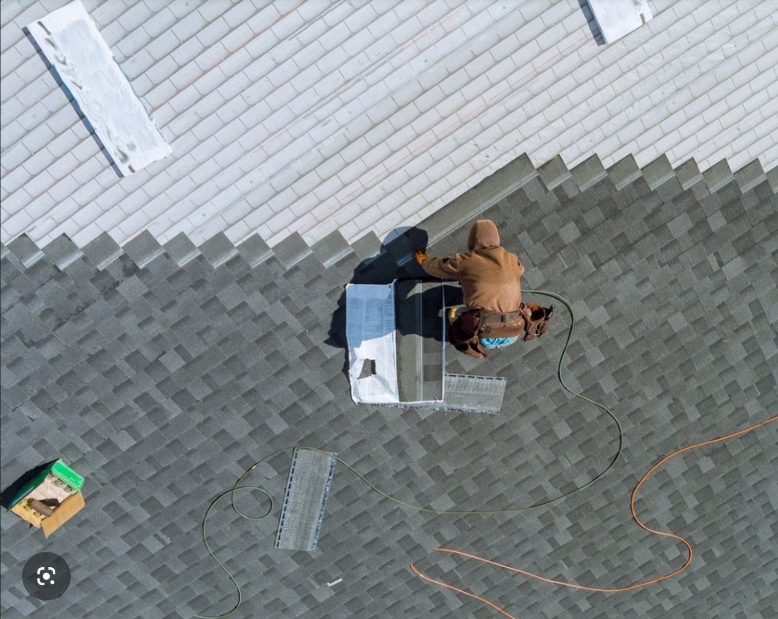 Overhead view of a person working on the roof, sitting on a ladder, wearing a hooded jacket, with tools and equipment nearby.