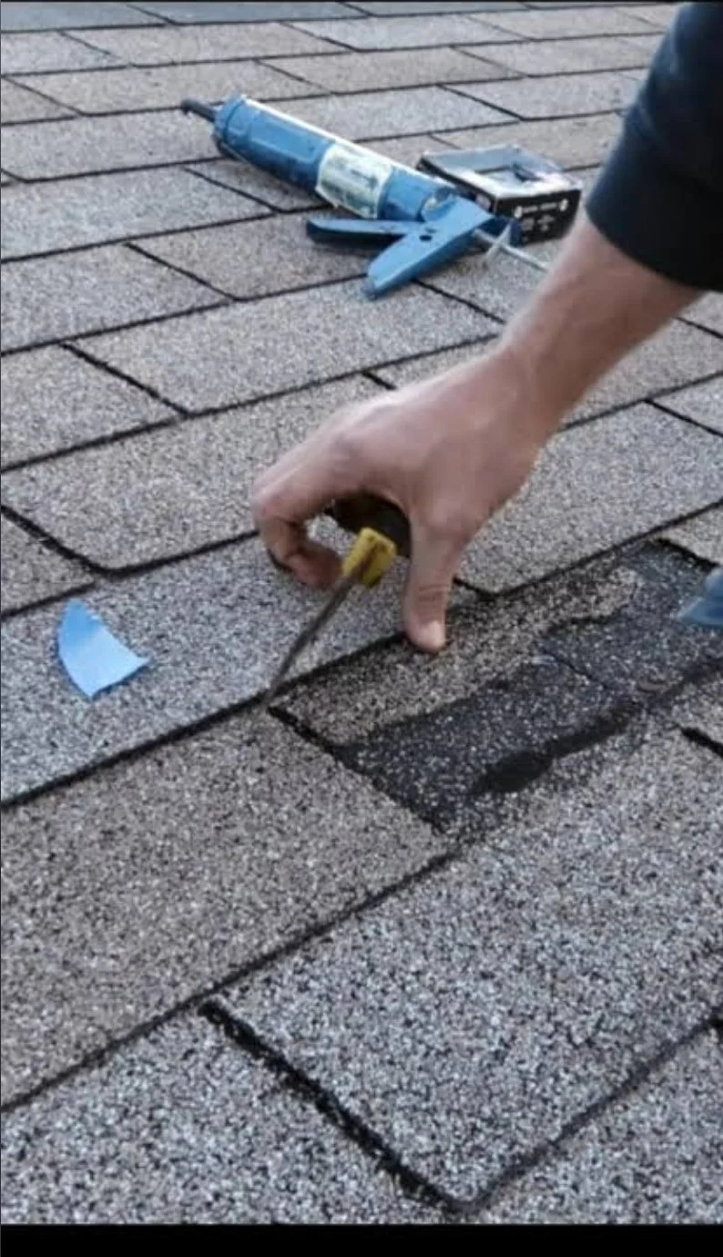 A person is using a screwdriver to remove or repair a shingle on a roof with asphalt shingles, and there is a caulking gun and a small black box nearby.