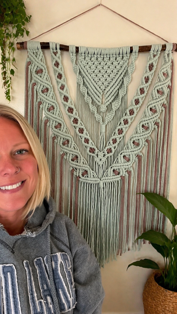 A smiling woman with blonde hair in a gray hoodie is standing in front of a large, decorative macrame wall hanging with intricate patterns and tassels, hung on a wall with a plant nearby.