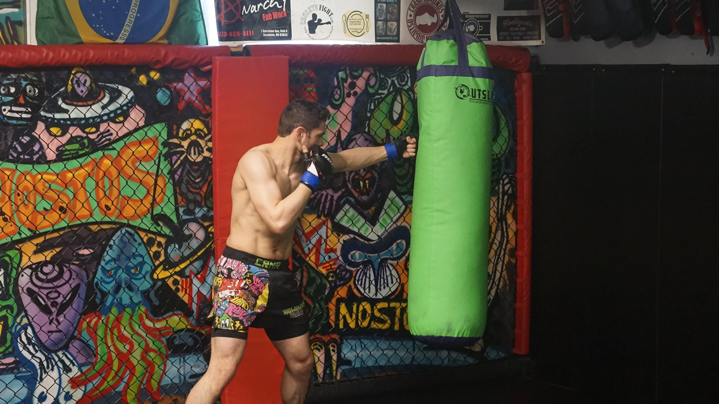 A shirtless man wearing colorful shorts practicing boxing in a gym, hitting a green punching bag.