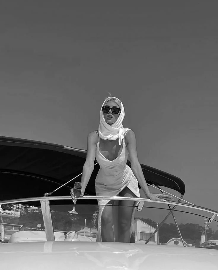 A woman in sunglasses and a headscarf standing on a boat, holding a glass of champagne against a clear sky.