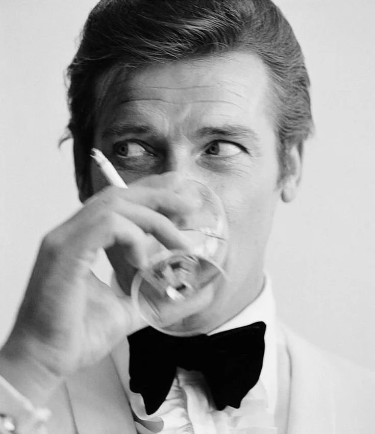 A black-and-white photo of a man in formal attire, wearing a bow tie, drinking from a glass, and holding a cigarette in his right hand, with hair neatly combed back, looking to the side.