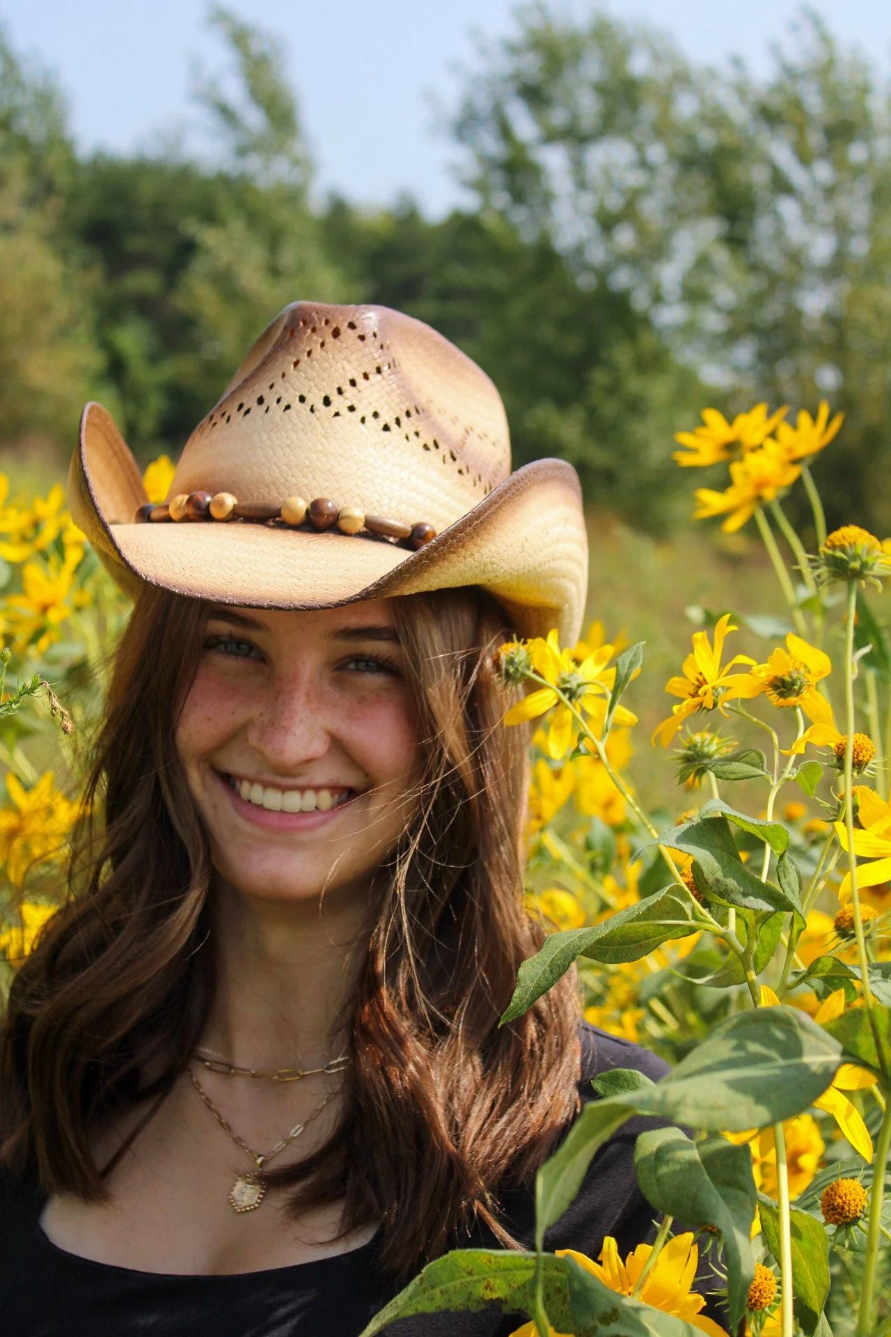 A woman wearing a cowboy hat and black shirt, smiling in a field of yellow flowers with green trees in the background.