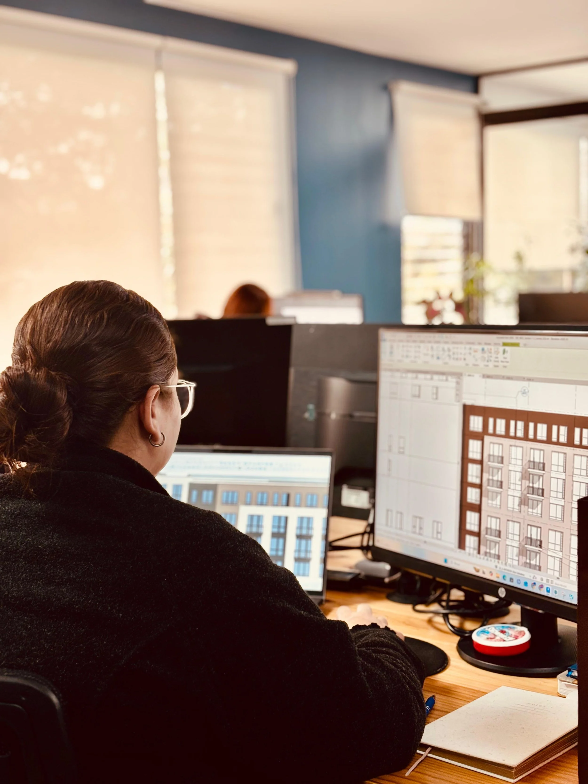 Office workspace with a woman working on a computer, surrounded by multiple monitors displaying architectural plans, on a wooden desk with a notebook, a pen, and a decorative container.
