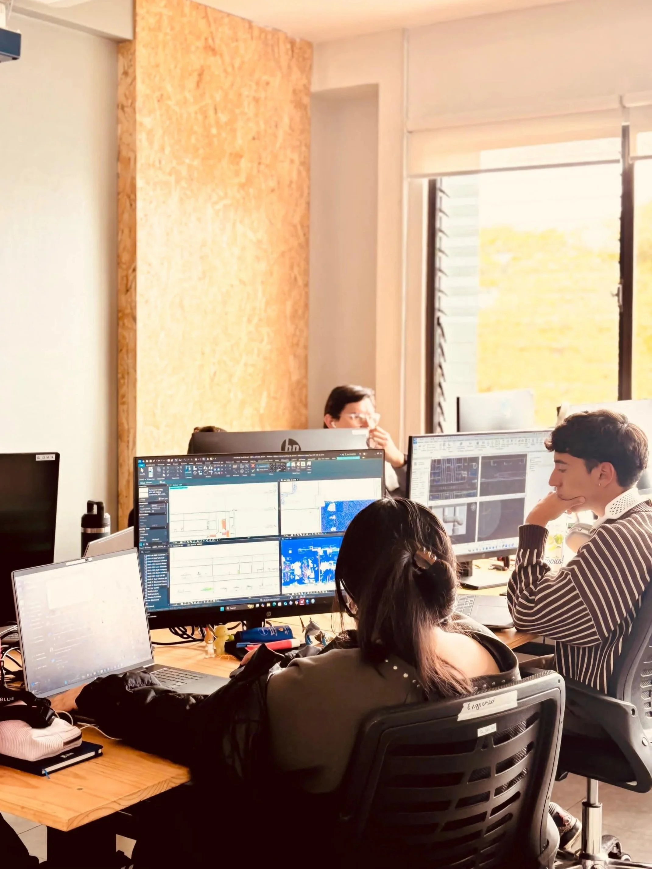 Office workspace with multiple people working at computers with large monitors, some people wearing glasses, a brick wall, and a window providing natural sunlight.