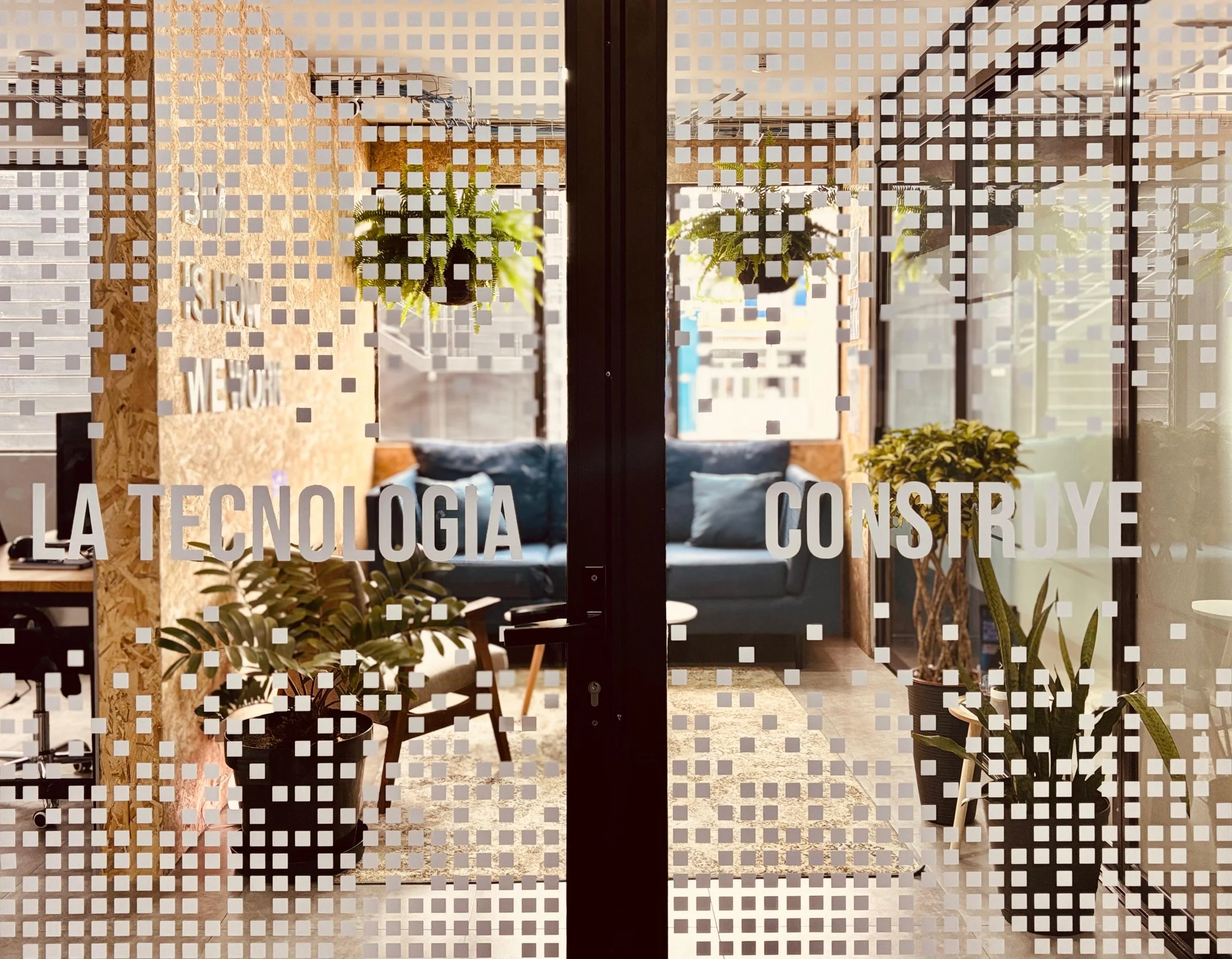Interior view through glass door with a pattern of white squares, showing potted plants, a seating area with cushions, and a textured wooden wall with part of a sign.