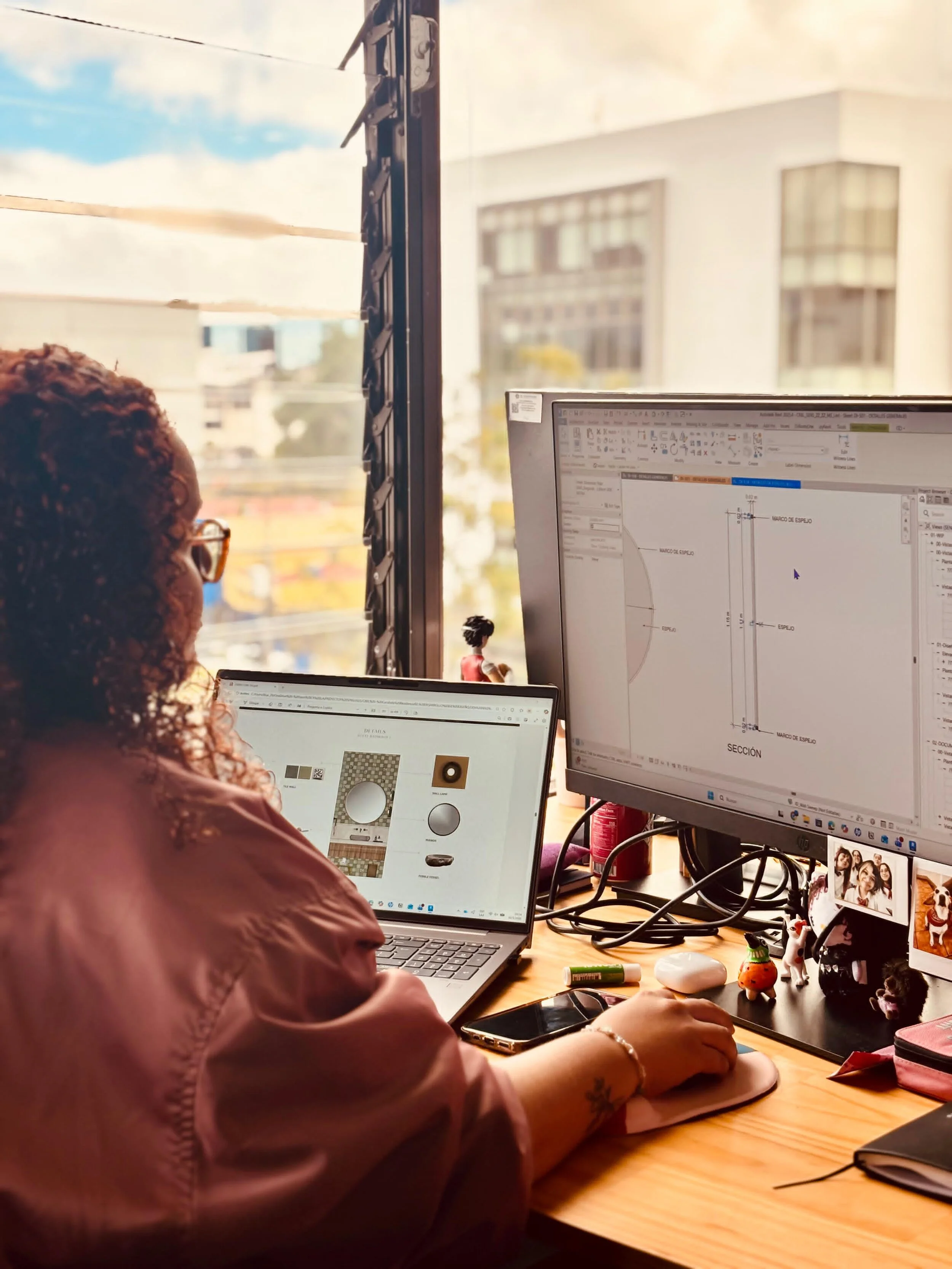 Person working at a desk with a laptop and dual monitors, one displaying design software, in a bright office with large windows.