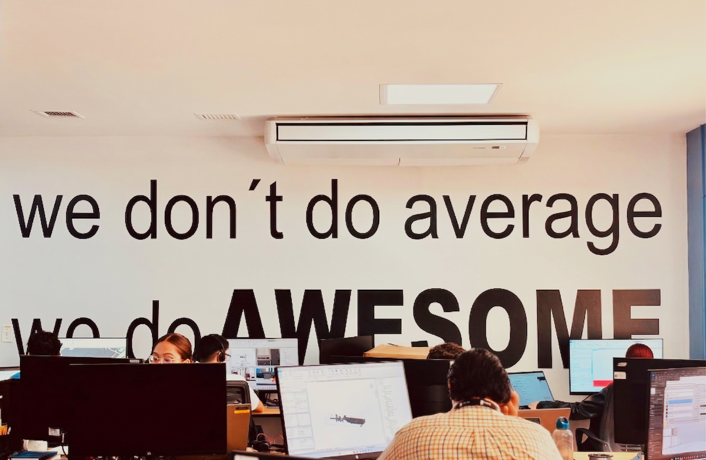 Office space with several employees working at desks with computer monitors. A large wall in the background has text that reads 'we don’t do average' and 'who do AWESOME'.