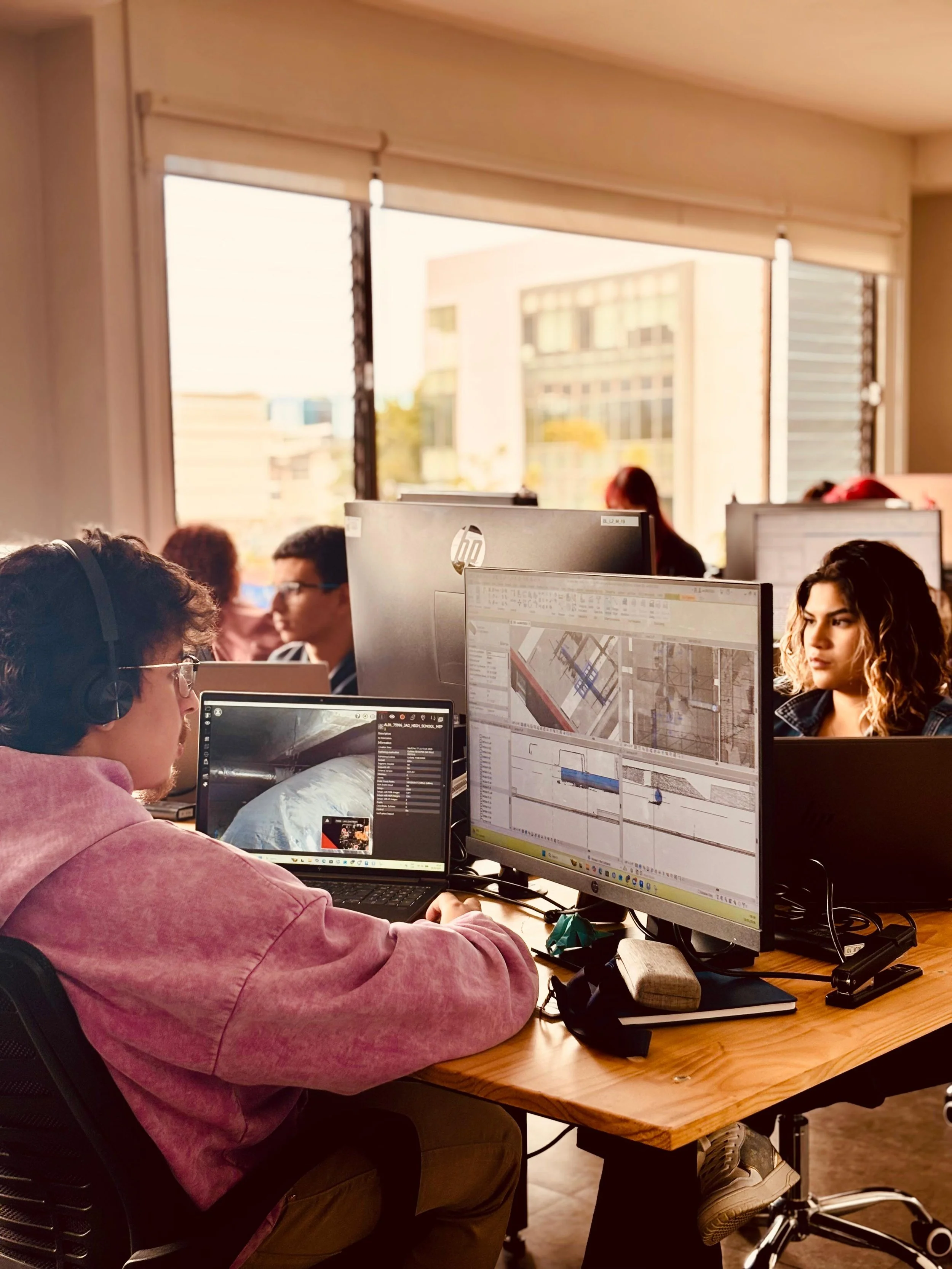 Office scene with people working on computers, large windows with cityscape view, monitors displaying maps and design software.
